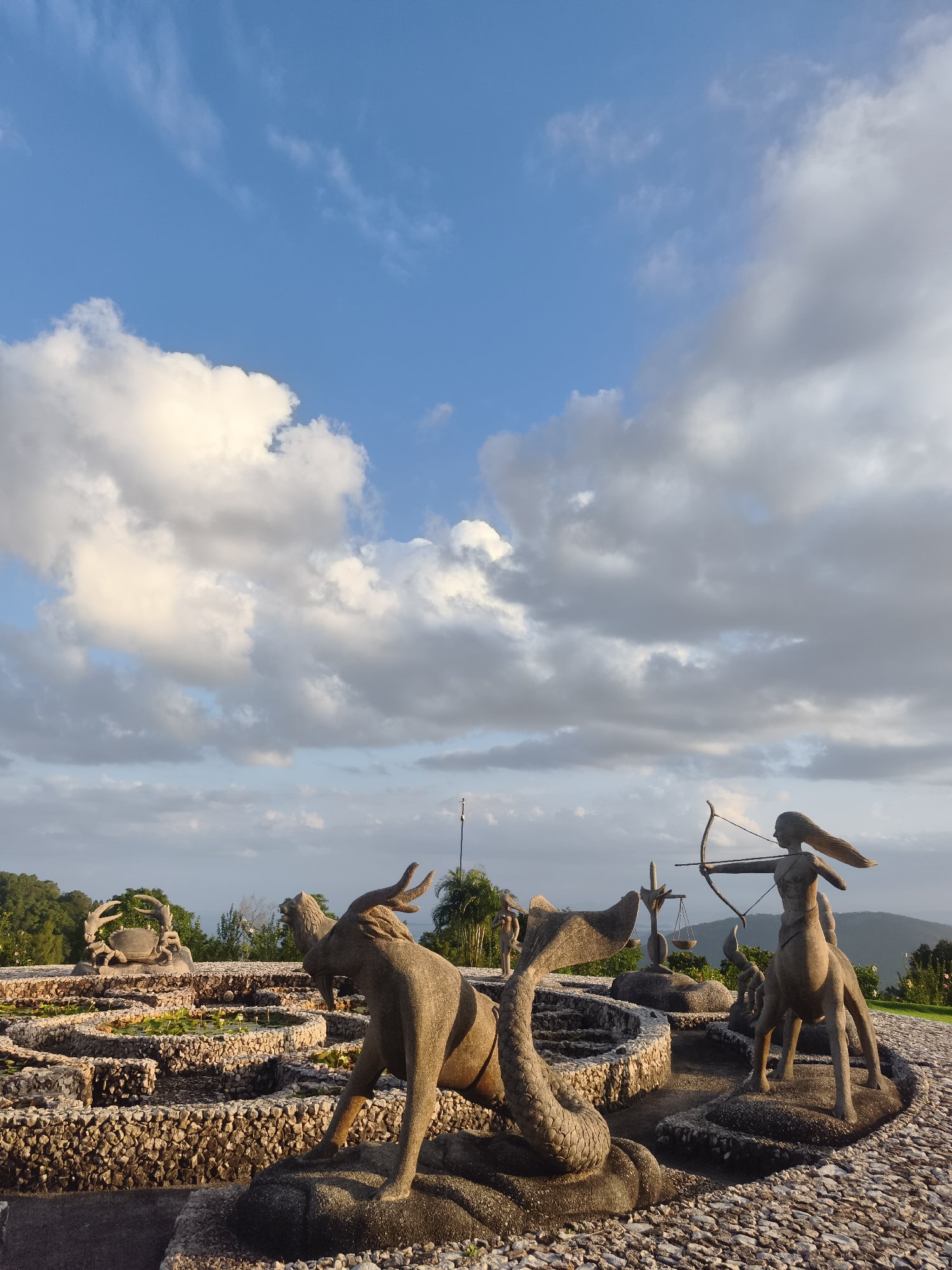 The image shows a stone sculpture garden featuring zodiac signs located at the Big Buddha Temple in Ko Samui District, Thailand. The sculptures depict various astrological symbols, including a centaur archer (Sagittarius), a mermaid-like creature with a goat's head (Capricorn), a crab (Cancer), and scales (Libra). These sculptures are arranged around a series of pools and stone pathways, with lush green hills and a body of water visible in the background under a sky with scattered clouds. The sun appears to be setting, casting a warm light on the scene.
