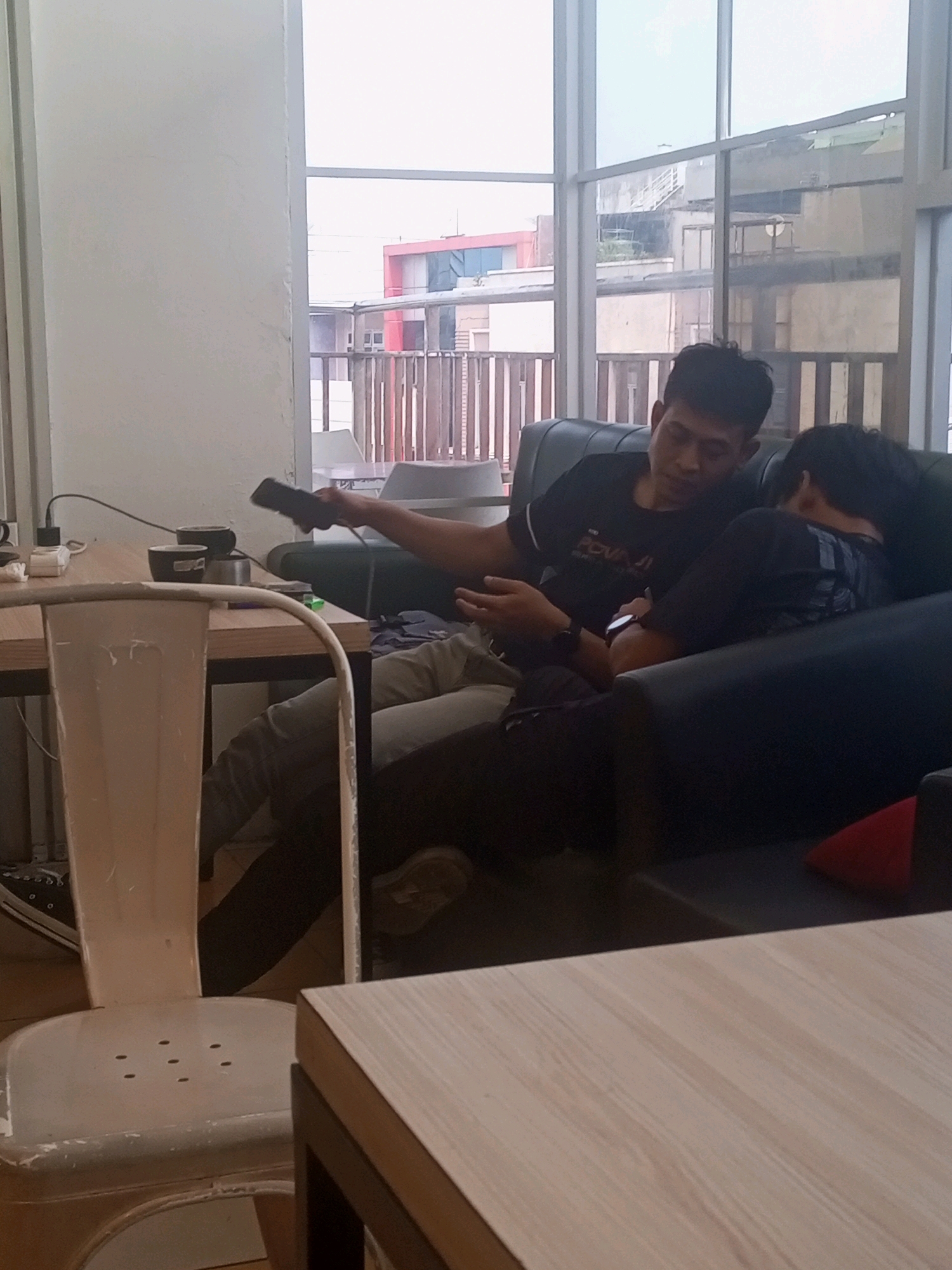 The image depicts an indoor scene in Bandar Lampung, Indonesia, featuring two individuals seated on a dark couch. One person, appearing to be a male, is wearing a black t-shirt and is actively engaged with a mobile phone, holding it in their right hand. This individual is looking towards the phone. The second person, lying down with their head on the couch, is wearing a black t-shirt with a graphic print. The environment includes a large window with a view of urban buildings, suggesting the location is likely on an upper floor of a building. A light-colored chair is partially visible in the foreground, and a wooden table with two black cups and charging cables is present to the left. The overall lighting suggests an overcast or indoor environment.