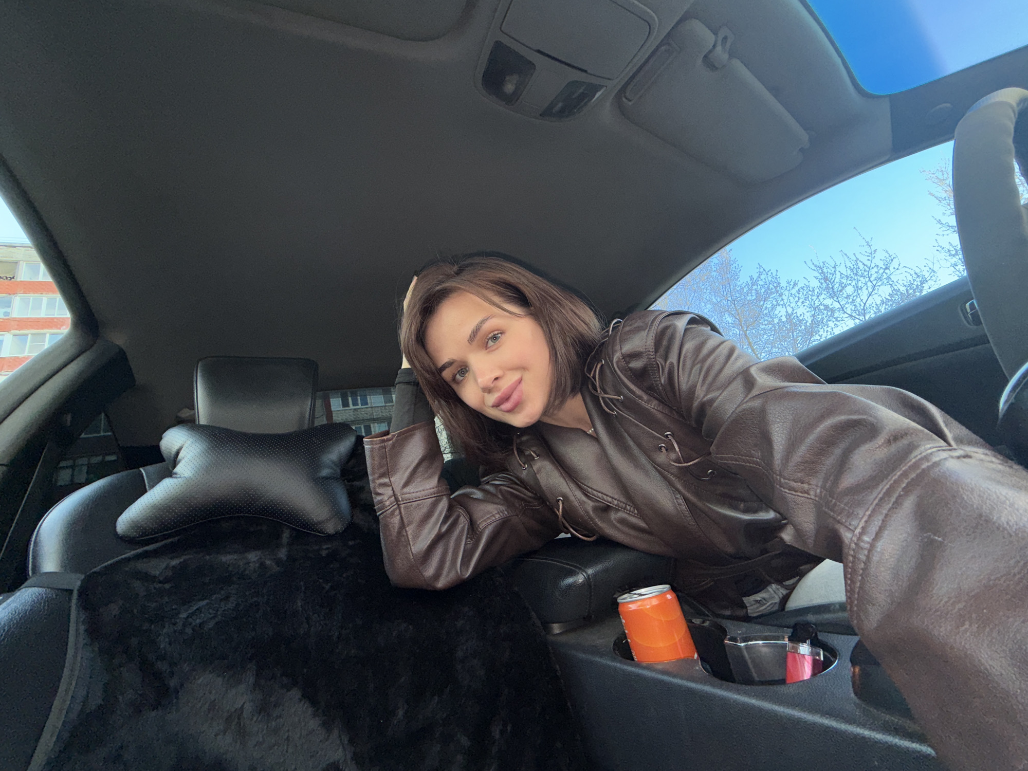 A young woman poses inside a car. She is fair-skinned with short brown hair and is wearing a brown leather jacket. She is leaning on the center console of the car, and an orange soda can is visible in one of the cupholders. The car's interior is dark gray. A black seat cover is visible on the driver's seat. Outside the car windows, a clear blue sky and bare trees can be seen, as well as a residential building in the background. Based on architectural style, building materials, and vegetation, the location is consistent with Perm, Russia.