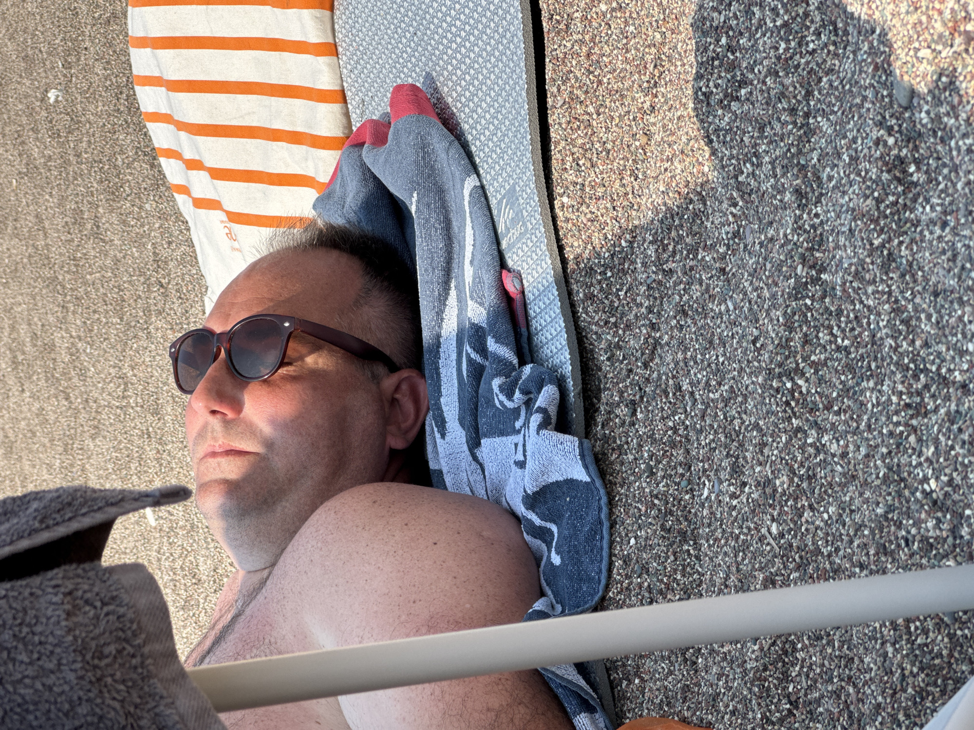 A man is lying on a beach in Boreti, Montenegro, on a sunny day. He is wearing sunglasses and appears to be asleep, resting on a blue and white patterned towel that is placed on a gray mat. To his right, an orange and white striped towel is visible. He is partially covered by a gray towel draped from above. The beach is covered in small pebbles. A white pole is visible in the foreground, likely part of a beach umbrella or chair, casting a shadow on the ground. The scene suggests a relaxed and leisurely atmosphere typical of a beach vacation.