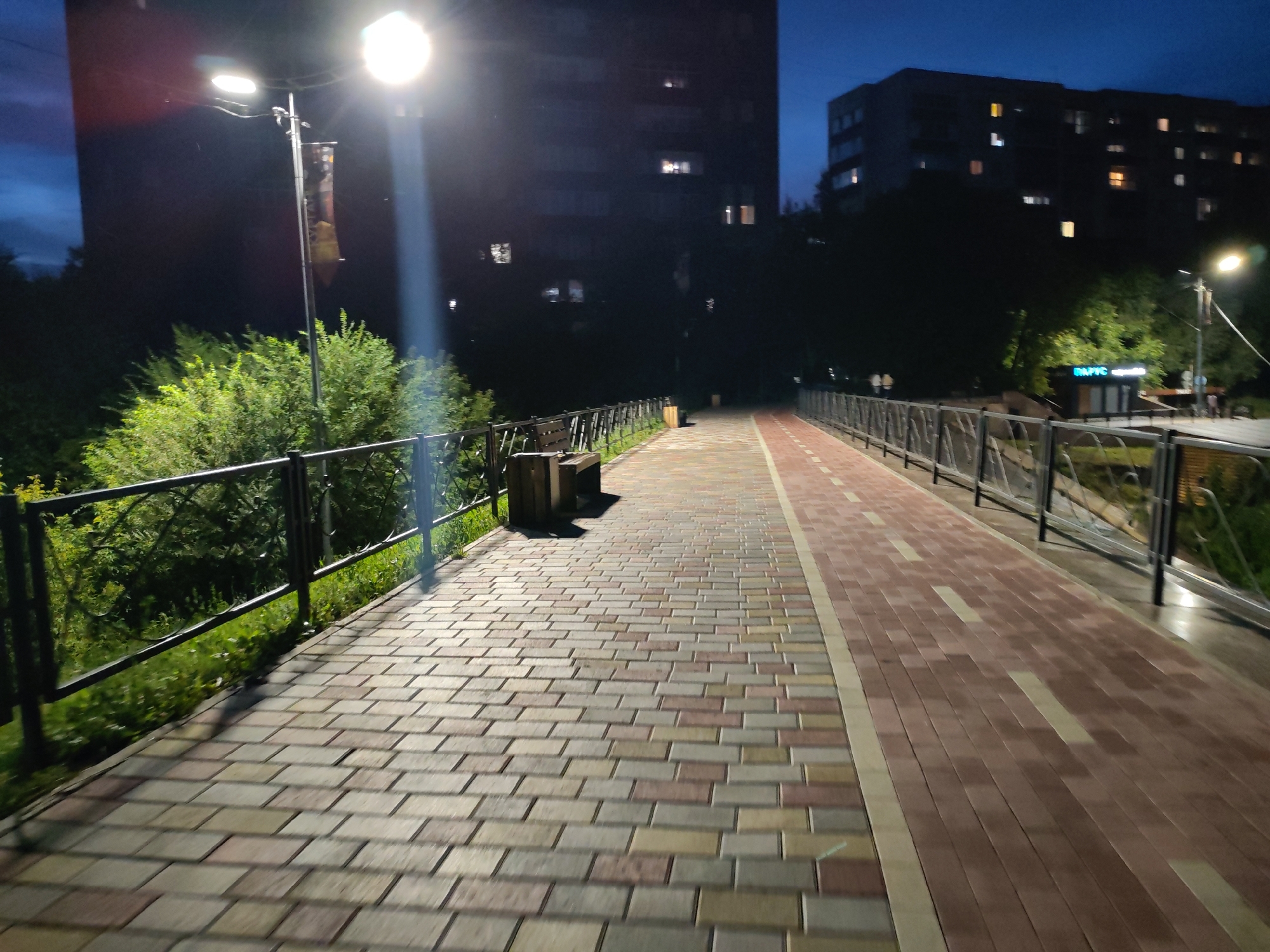 The image depicts a pedestrian walkway at night in an urban environment. The walkway is paved with interlocking bricks in various shades of beige and brown, with a separate red-bricked lane for cycling or jogging. A dark metal railing runs along the side of the walkway, separating it from a grassy area with trees and bushes. In the background, two multi-story buildings are visible, with lights on in some of the windows. A street lamp illuminates a portion of the walkway. A wooden bench and a trash receptacle are also present. The sky is a deep blue, indicating it is either dusk or early dawn.