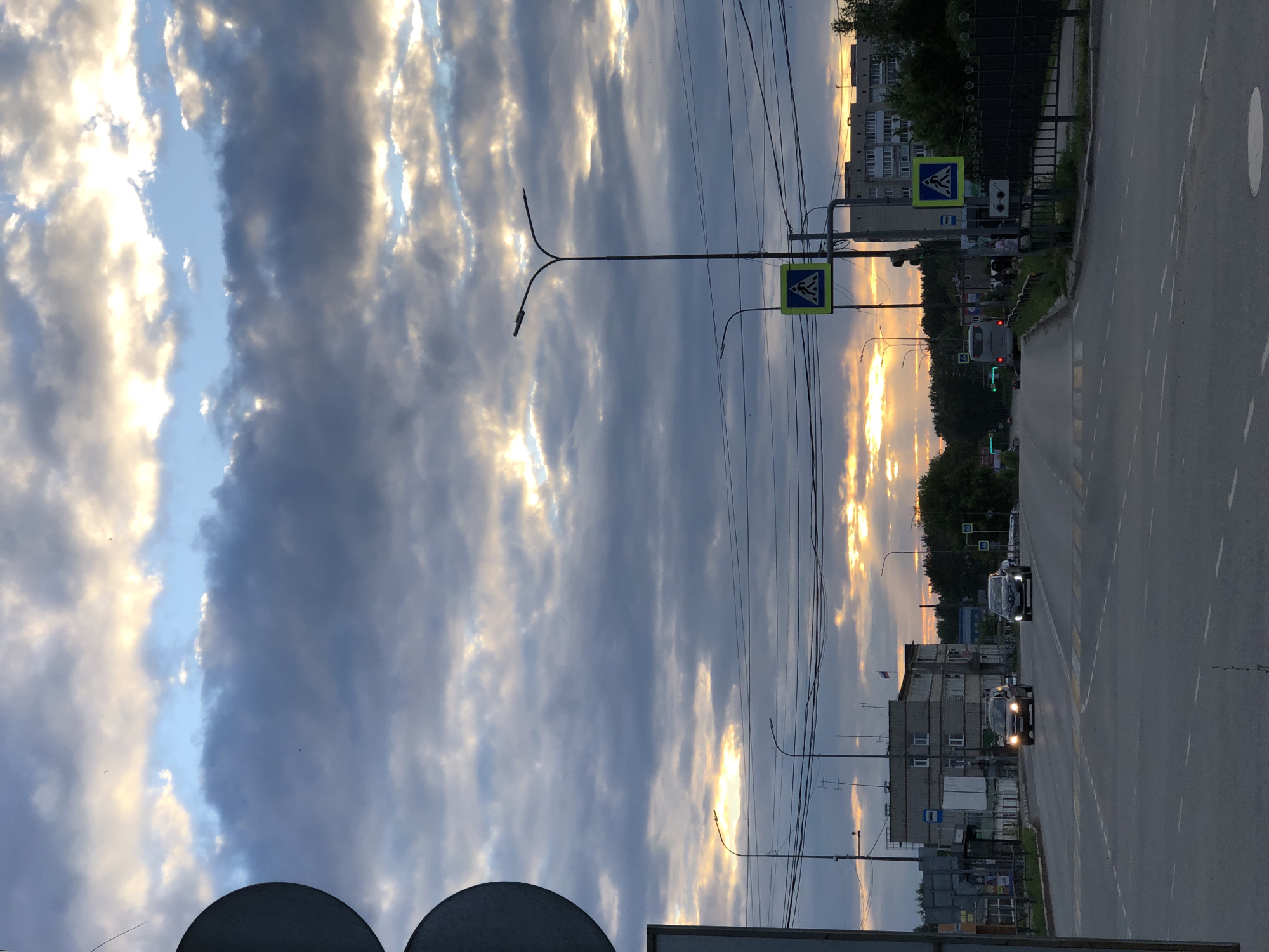 The image depicts a view of a city street in Chaikovsky, Russia, at dusk. The sky is filled with dramatic clouds, with patches of golden light from the setting sun breaking through. The foreground shows a wide, paved road with white lane markings and pedestrian crossings. Several cars are visible on the road, including a dark-colored SUV and a silver sedan traveling towards the viewer.
On the right side of the road, there are streetlights with visible lamps. A yellow pedestrian crossing sign is mounted on a pole. Further down the road, apartment buildings of several stories can be seen, with some displaying flags. Trees line the side of the road in the distance. Electrical wires are strung overhead, supported by poles. A bus is also visible in the distance on the left side of the road. The overall scene suggests a typical urban environment during a transitional time of day.