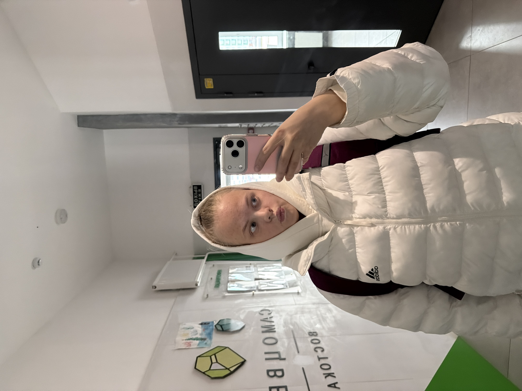 A young woman is taking a selfie in a hallway. She is wearing a white Adidas puffer jacket with a hood and a burgundy backpack. She is holding a pink iPhone in her right hand. The hallway is white with a gray concrete ceiling. There is a black door in the background. A transparent display case is visible with green and blue geometric shapes in front of the display. The sign, "ВЕЧЕРНИЙ ИЖЕВСК" (Evening Izhevsk), is written in Russian. The location is Izhevsk, Russia.