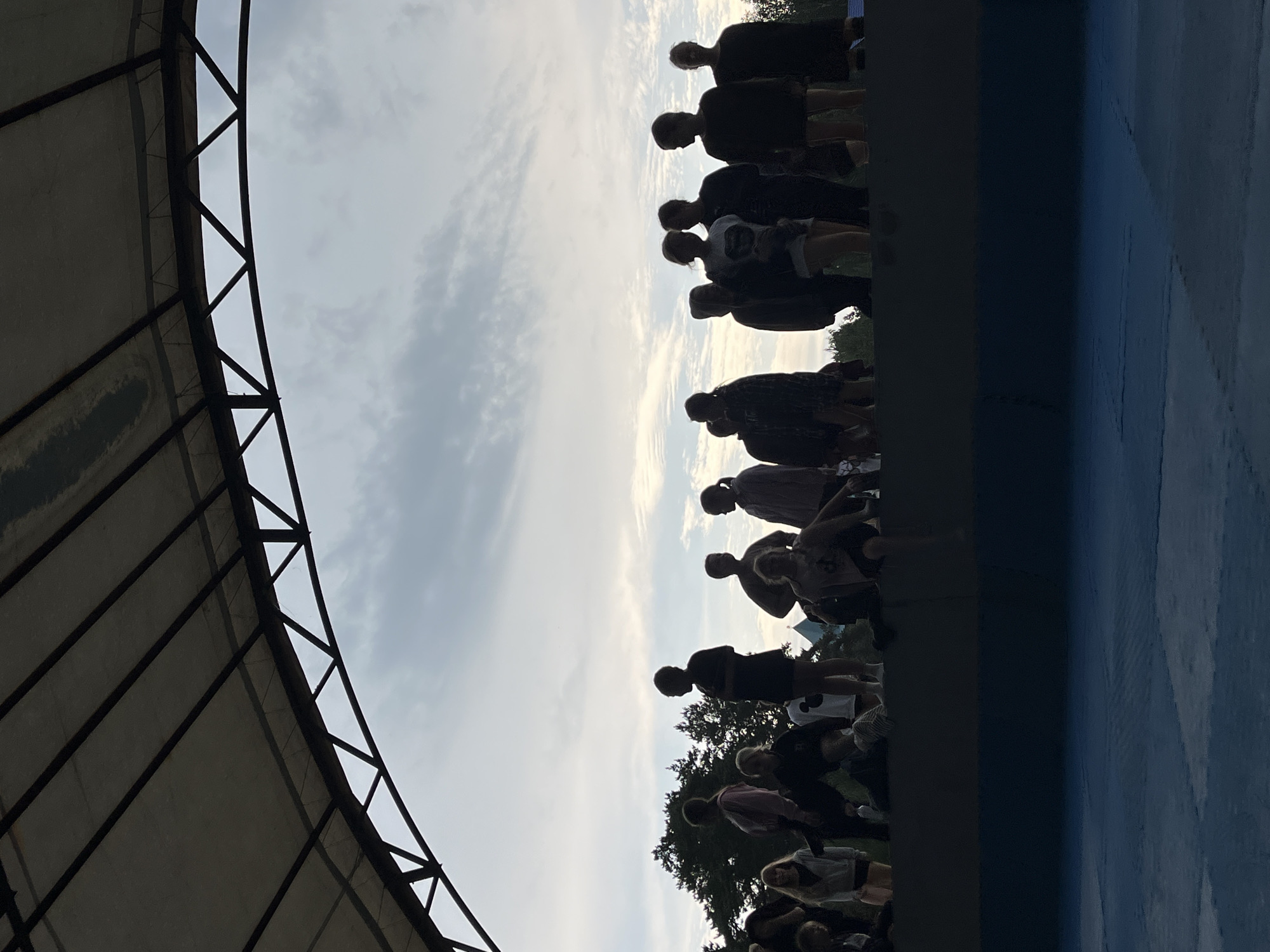 The image shows a group of people silhouetted against a bright, cloudy sky. They appear to be standing on a raised platform or tiered seating area, possibly for an outdoor event. To the left, a portion of a large, dark canopy with a metal frame is visible, suggesting a covered stage or seating area. The people are facing towards the right of the frame. The time of day appears to be late afternoon or early evening, as indicated by the quality of light and the long shadows. The weather seems to be partly cloudy. The overall atmosphere is somewhat unclear due to the silhouettes, but the gathering of people implies a communal activity or event. There is no visible text in the image.