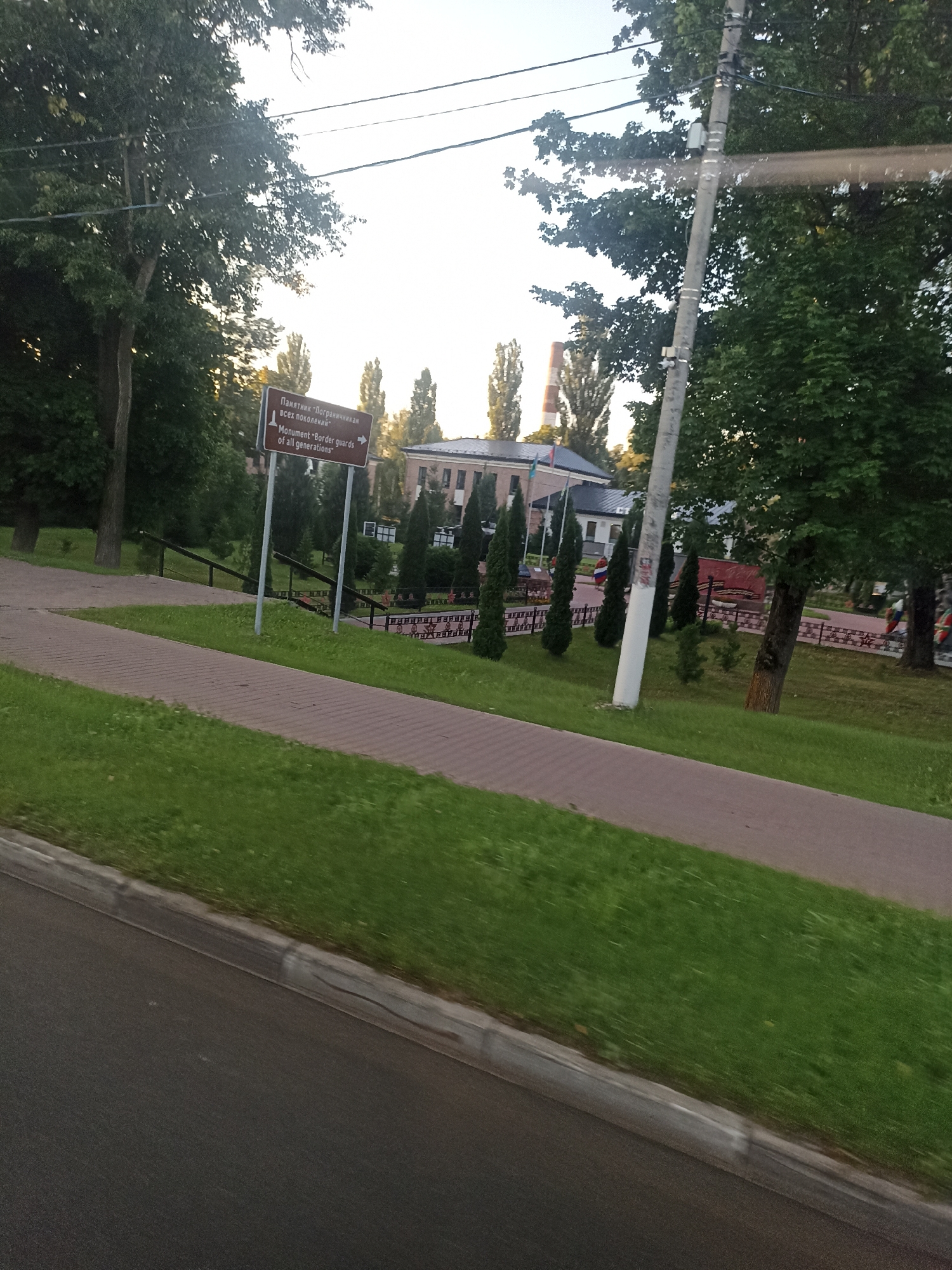 The image shows a brown directional sign with text in Russian and English. The Russian text reads "Памятник "Пограничникам всех поколений"", and the English text reads "Monument "Border guards of all generations"". An arrow points to the right, indicating the direction to the monument. The sign is positioned next to a paved walkway and a grassy area, with trees and buildings in the background. The scene appears to be outdoors, possibly in a park or public space within the city of Chekhov, Russia. The lighting suggests it is daytime.