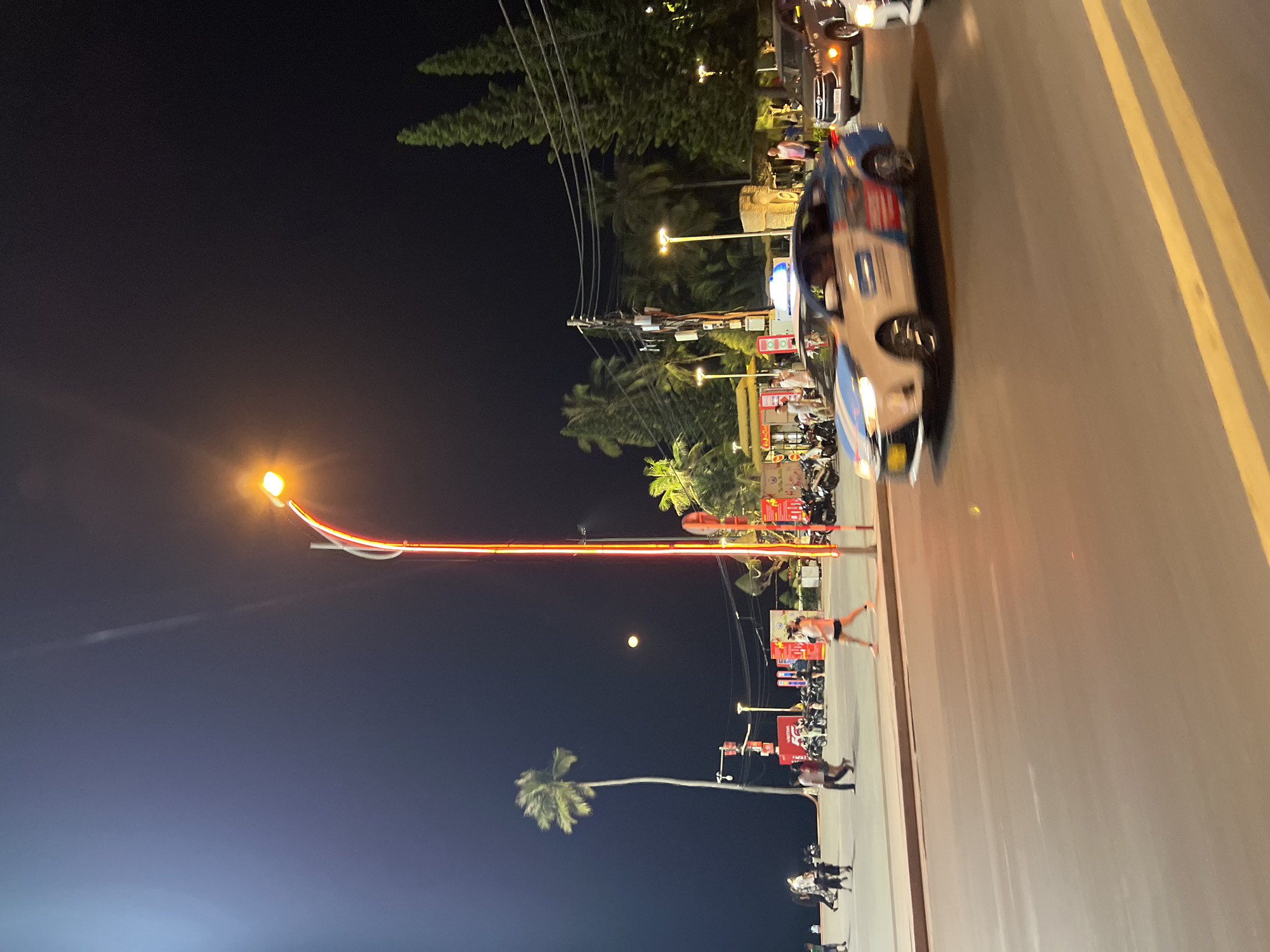 At night, a street scene in Nha Trang, Vietnam, shows a street with a car, various palm trees, streetlights, and people. The road has yellow lines and several parked scooters are on the left side. The scene is illuminated by streetlights. The sky is dark with a visible moon. A tall palm tree is positioned in the bottom left corner, and several other palm trees can be seen farther down the street. People are visible, mostly walking alongside the road. Streetlights with arched designs are visible. A car is visible and partially blocking the lane.