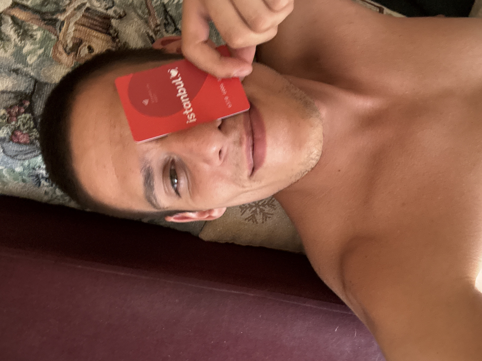A young man, with short brown hair and light brown eyes, is lying down on a bed or couch. He is holding a red card, partially covering his left eye, which has the text "istanbul" printed on it in white lettering. The man is shirtless and appears to be in a bedroom. The background shows a patterned fabric, possibly a blanket or upholstery. No specific landmarks or indications of Sofia, Bulgaria are visible in the image.