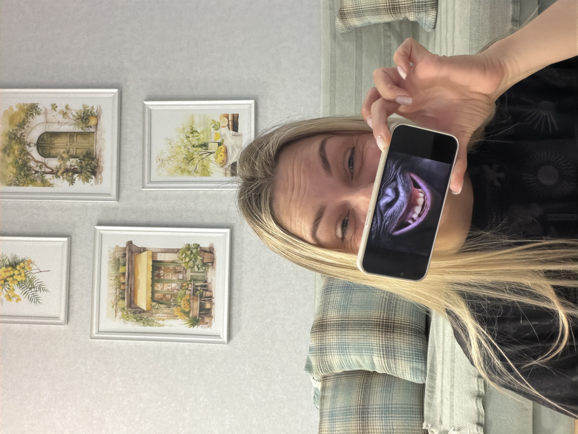 An indoor scene features a blonde woman holding a smartphone, slightly obscuring her face, in what appears to be a living room or residential setting in Votkinsk, Russia. The image is oriented vertically with the woman on the right side of the frame.
The woman has long, straight blonde hair and is wearing a dark-colored top. Her visible eye is open and looks towards the camera. She holds a white-cased smartphone in her right hand, positioned in front of her face. The screen of the phone displays a close-up image of a gorilla's face, specifically its mouth open in a wide grin or snarl, revealing teeth. Her forehead shows some subtle wrinkles, suggesting a possible expression of mild effort or amusement as she holds the phone.
The background consists of a light-colored, possibly off-white or light grey, wall with a subtle textured pattern. Four framed pictures hang on the wall, arranged in two rows of two. The artworks depict serene, watercolor-style scenes:
- Top left: An arched doorway surrounded by lush green foliage and yellow fruits, possibly lemons.
- Top right: A still life composition featuring bottles, fruit, and greenery on a table.
- Bottom left: A bouquet of delicate yellow flowers, resembling mimosa, with green leaves.
- Bottom right: A quaint storefront or cafe facade with a yellow awning and various potted plants.
Part of a sofa or armchair is visible, covered with plaid and striped cushions or blankets in shades of blue, grey, and cream, providing a cozy backdrop. There is no visible text in the image. The time of day is not discernible, as the lighting appears to be artificial indoor lighting.