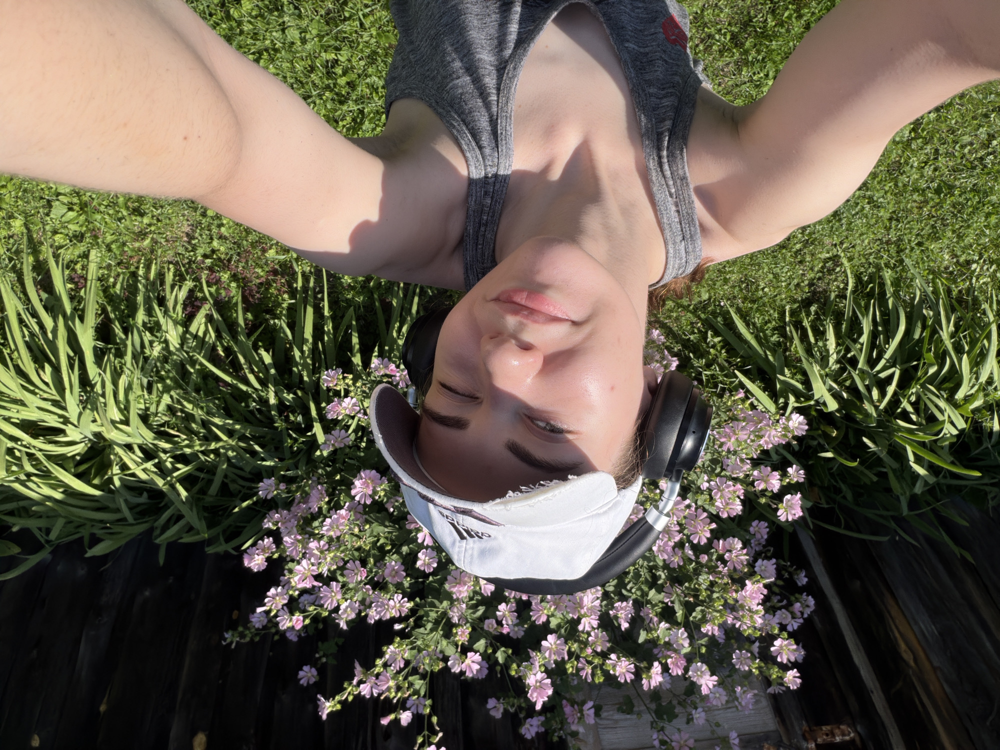 A young adult, identified as female by her facial features and attire, is depicted in a selfie taken outdoors. She is wearing a white baseball cap with a distressed brim, black headphones, and a grey tank top. Her right eye is partially closed in a wink, and her skin appears to be illuminated by natural sunlight, suggesting it's daytime. The background features a dense cluster of pink flowers, a wooden fence, and lush green grass, indicating a garden or park setting. The lighting suggests a sunny day. Based on the provided metadata, this scene is located in Zavodoukovsk, Russia.