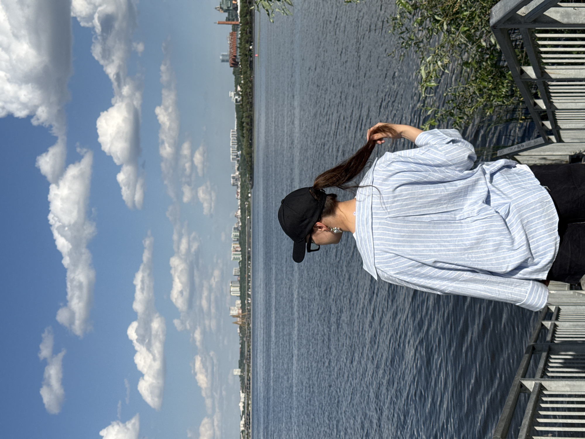 A woman stands outdoors, facing away from the camera, beside a body of water in Kazan, Russia. She wears a blue and white striped shirt, black pants, and a black baseball cap. Her brown hair is pulled back in a ponytail. The background shows the city skyline of Kazan with buildings and trees lining the opposite side of the water. The sky is blue with puffy white clouds. A metal railing is visible on the right side of the image.