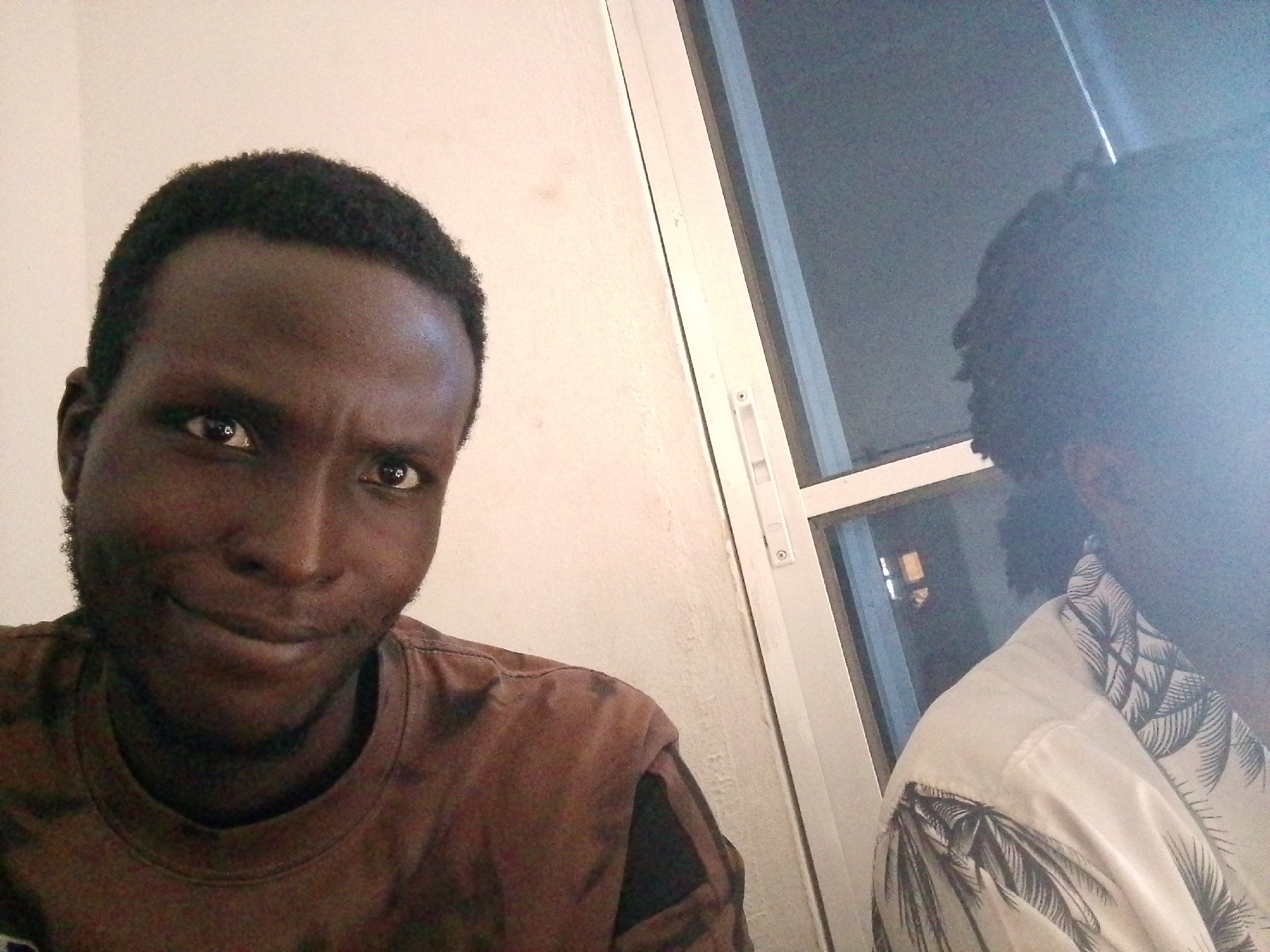 Two people are visible in this close-up, self-taken photo. The person in the foreground, on the left, is a young Black man with short, dark hair and a light beard. He is looking directly at the camera with a slight smile, his head slightly tilted. He is wearing a brown t-shirt. Behind him, to the right, is another person whose face is largely obscured by glare and focus. They appear to be wearing a white shirt with a dark, tropical leaf pattern. The background is divided by a white doorframe or window frame. To the left of the frame, the wall is a light, neutral color. To the right, through the frame, is a darker area that appears to be a window with interior lights visible, suggesting it is nighttime or a dimly lit interior. The overall lighting is uneven, with a strong light source on the left casting shadows. The image appears to be taken indoors. There is no visible text in the image.