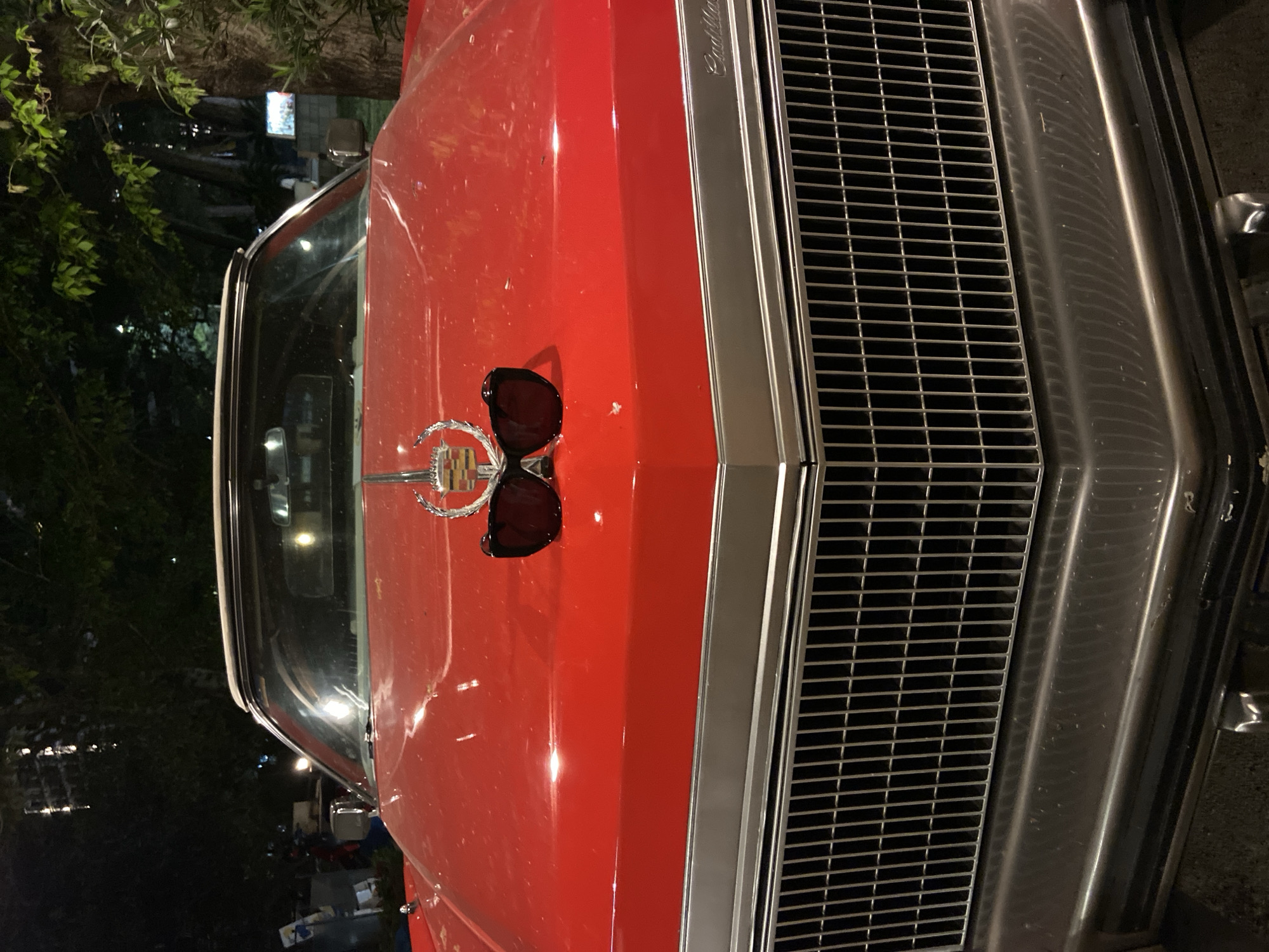 The image shows the front hood and grill of a red Cadillac in a somewhat dark environment. A pair of sunglasses rests on the hood near the Cadillac emblem. The background shows foliage and indistinct buildings. It's challenging to specifically locate the image within Budva, Montenegro, without additional information about the environment.