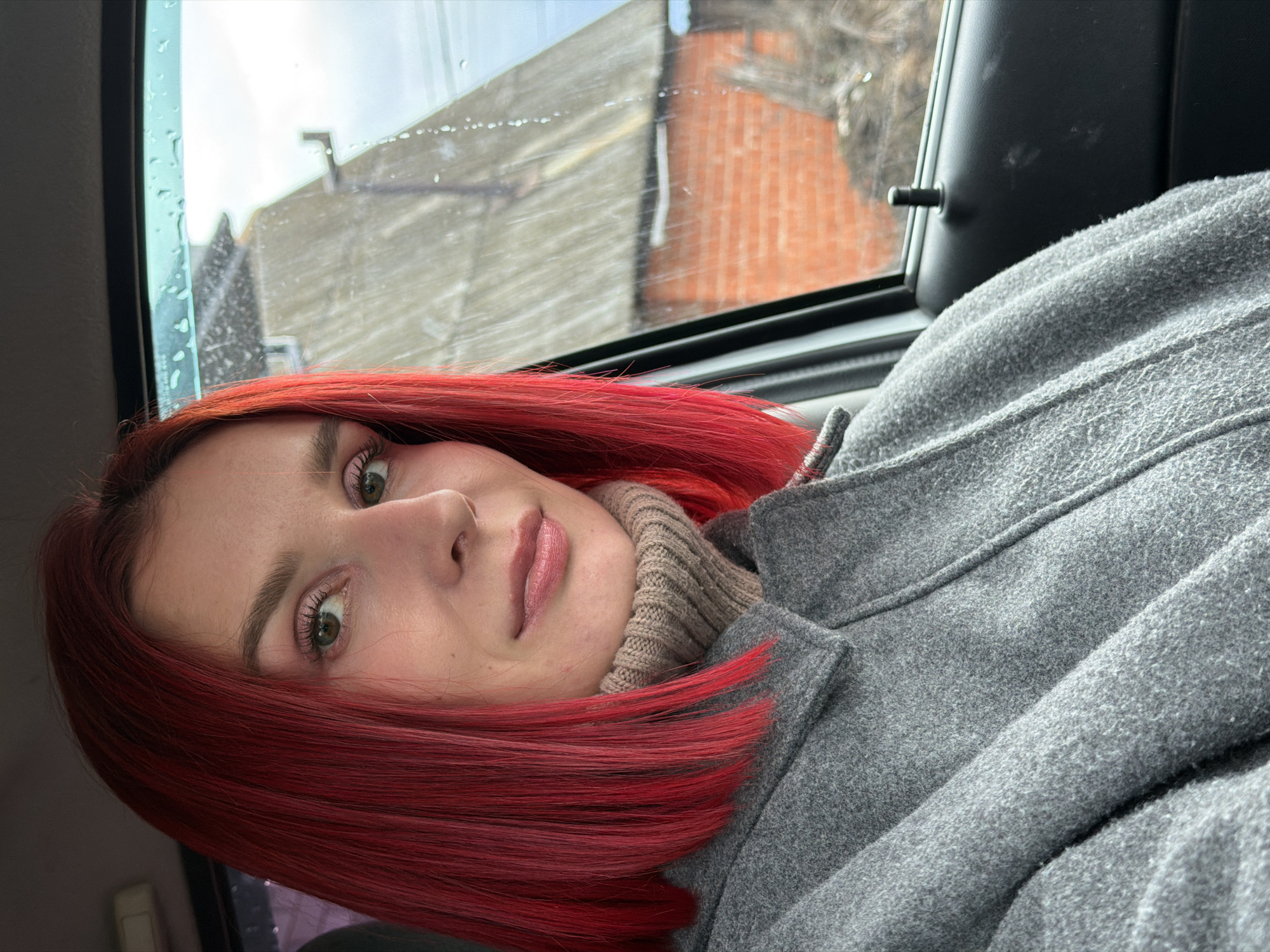 A young woman with vibrant, shoulder-length red hair is captured in a close-up, selfie-style photograph. She has fair skin, green or hazel eyes, and a neutral expression with subtly parted lips. She wears a warm, light grey, fuzzy-textured coat with a wide collar, and a beige or light brown ribbed turtleneck sweater underneath.
The woman is seated inside a vehicle, likely a car, with a window positioned to her left. The interior includes a black car door frame and a door lock mechanism. Through the window, an urban or suburban landscape is visible, featuring buildings with reddish-brown brickwork and lighter-colored facades, along with hints of bare tree branches or utility lines. The window glass is dotted with water droplets, suggesting recent rain, condensation, or ongoing damp weather. The sky appears overcast, providing soft, diffused lighting.
The activity taking place is a direct portrait or selfie, as the woman looks calmly into the camera. The time of day is daytime, evidenced by the ambient light. No discernible text is visible in the image. The overall setting is consistent with a cooler climate, aligning with the provided location context of Chaikovsky, Russia.