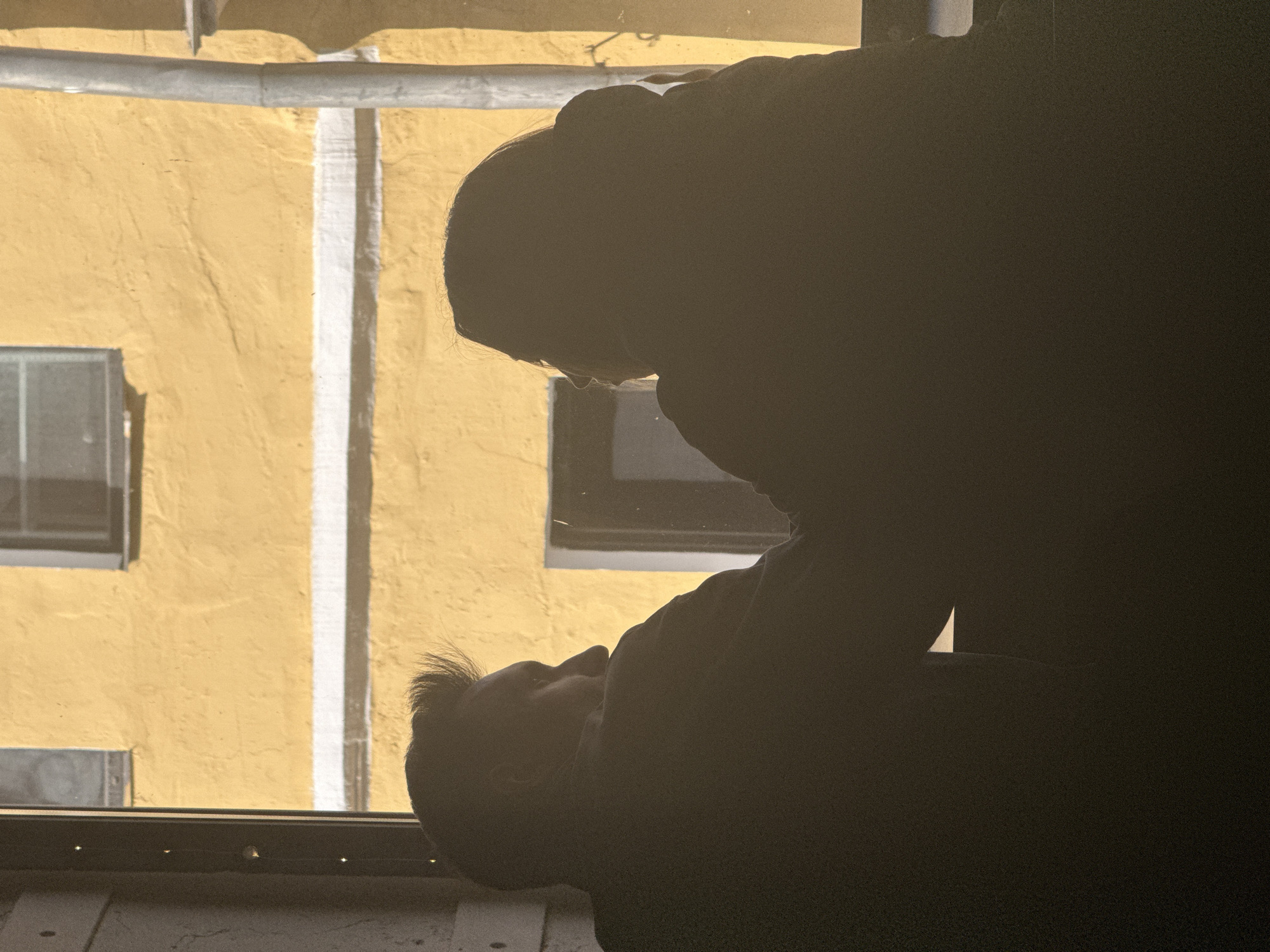 The image depicts two shadowed figures inside a building, appearing near a window. Outside the window, there is a view of a building with a yellow facade and white accents. The location appears to be in a city, potentially Moscow, Russia, based on the architectural style. The interaction between the figures and the environment is the subject of the image.