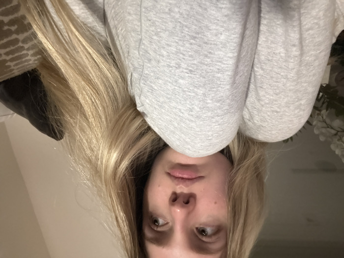 A young Caucasian female is depicted indoors in a close-up, chest-up portrait, situated within the city of Coral Gables, United States of America. She possesses long, straight, light blonde hair and light-colored eyes. Her head is tilted slightly, with her gaze directed upwards and to her left, presenting a pensive expression with slightly parted lips. She is wearing a light gray, heathered long-sleeved garment. Her right knee is drawn upwards, prominent in the lower foreground, with her right arm resting upon it. The background is dimly lit and out of focus, showing a white floral arrangement to the left and dark and patterned soft furnishings to the right.