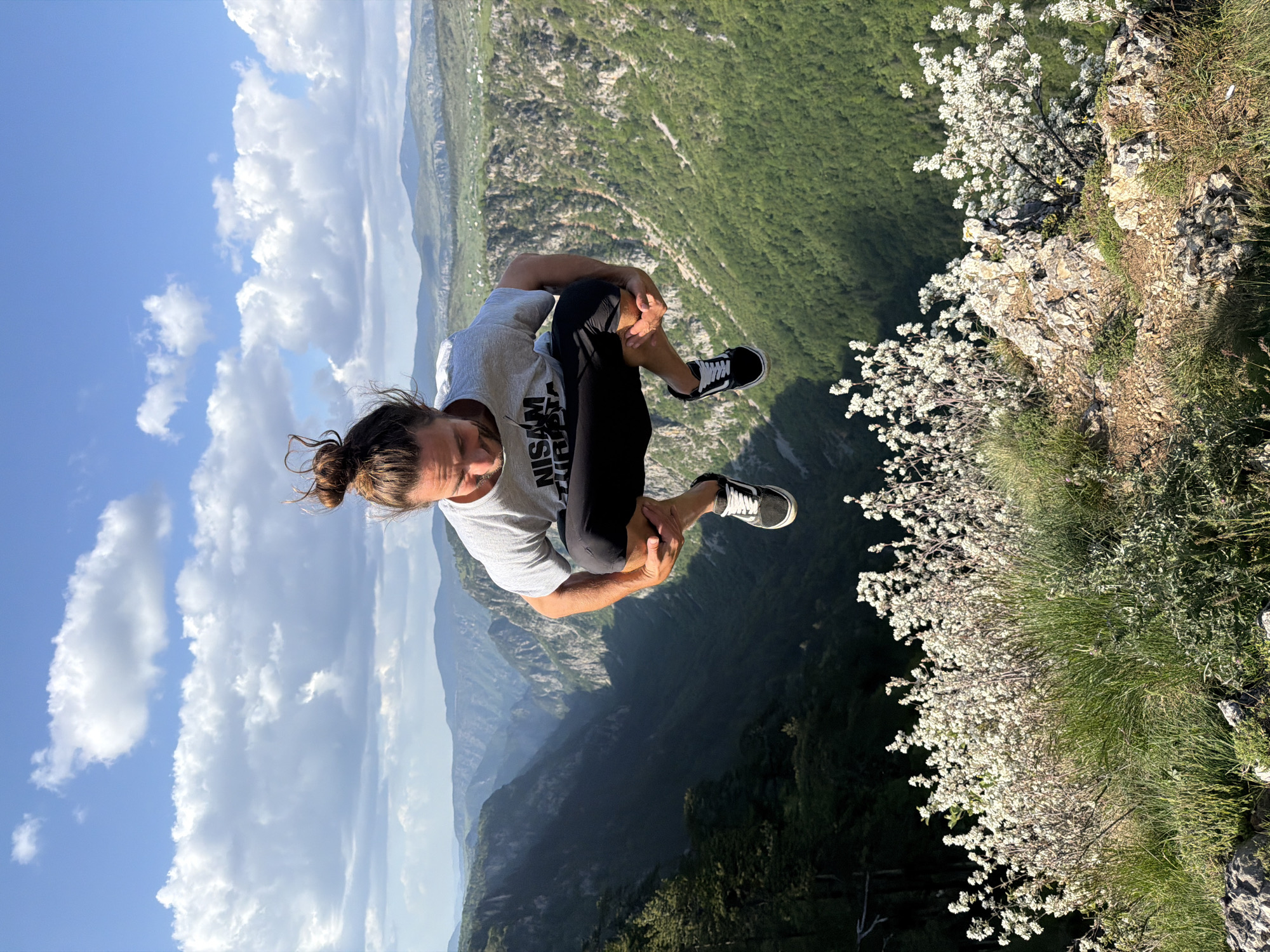 The image depicts a man in a crouched position on the edge of a cliff in Nedajno, Montenegro. He is fair-skinned with his hair pulled back in a bun and is wearing a grey t-shirt with black athletic pants and black and white sneakers. He is holding his knees, facing downward towards a steep drop-off into a mountainous valley. The background includes a blue sky with white clouds, a deep gorge, and a cliff edge with green vegetation and white flowers. The photo is taken from a perspective looking slightly upward towards the man.