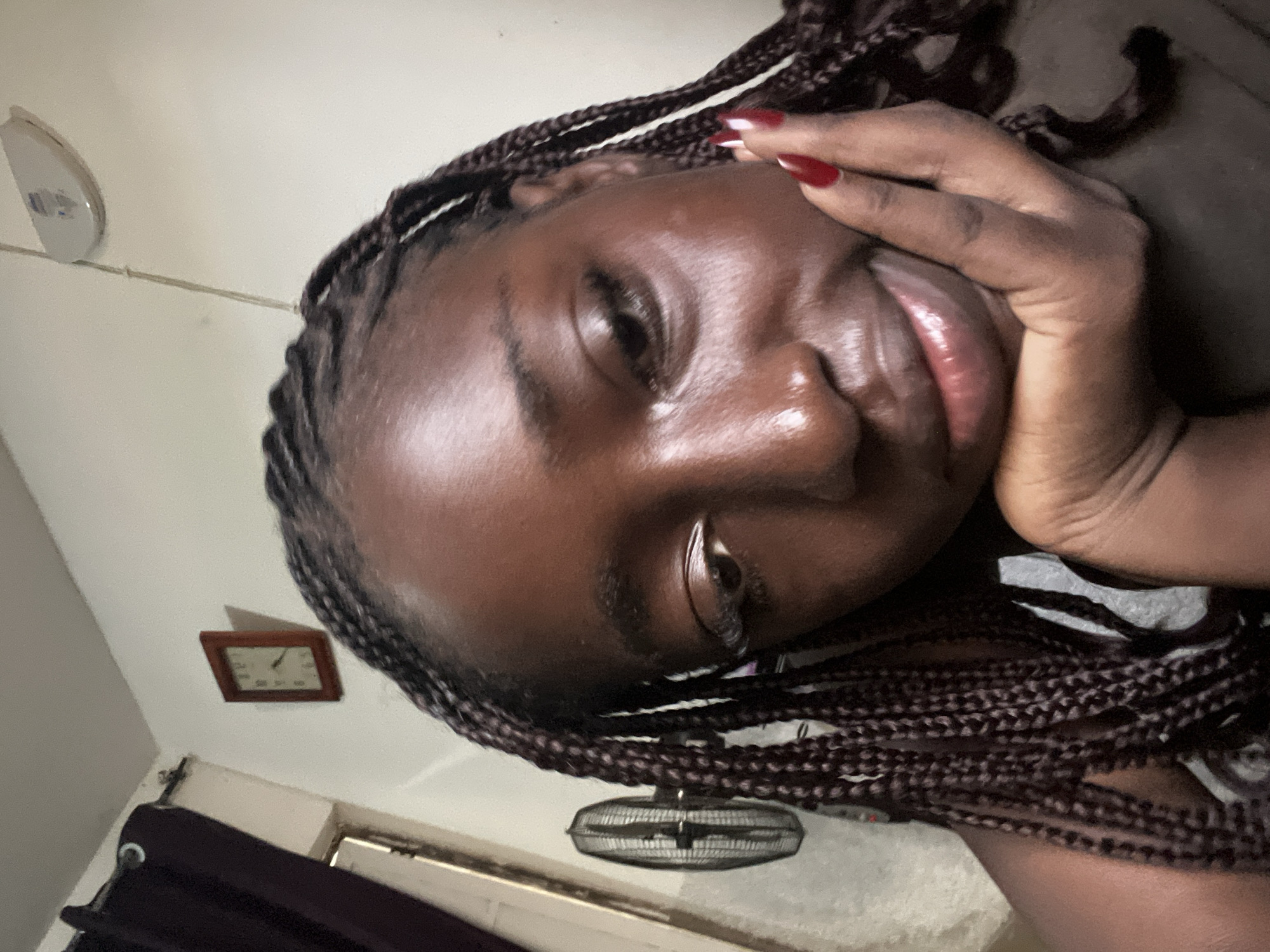 The image is a close-up selfie of a person with dark skin and braided hair, possibly a woman, lying down. Her face is angled towards the camera, with her hand partially covering her cheek. She has red nail polish. The background is a dimly lit room. On the wall behind her, there is a wall-mounted light fixture, a small, framed clock, and a small electric fan. The location in Abuja, Nigeria, cannot be determined from the image.