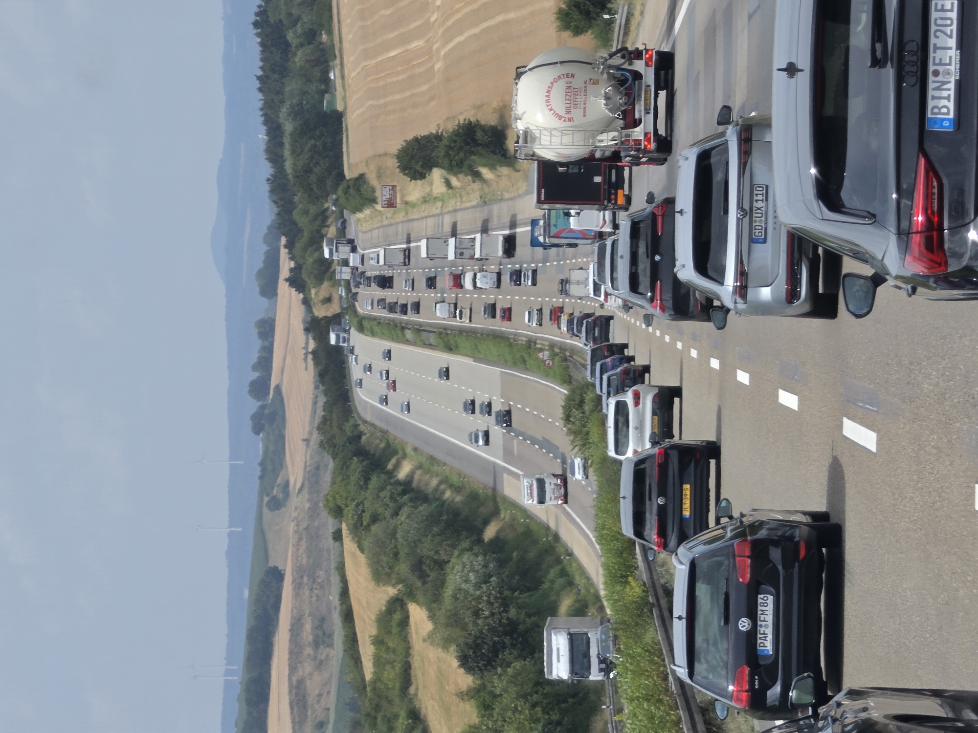The image shows a multi-lane highway in a rural landscape. The highway is congested with traffic, including cars, trucks, and a tanker trailer. The landscape is characterized by rolling hills with fields of crops and scattered trees. Wind turbines are visible in the distance on a hilltop. The sky is overcast. A black Volkswagen Golf with license plate "PAF FM 86" is in the foreground, to the left. A silver Volkswagen Passat with license plate "GD UX 110" is in the middle ground, to the right. A tanker trailer with the text "NILLEZEN EFFELT" and website "WWW.NILLEZEN.NL" is also visible. The image is captured from a slightly elevated perspective, looking down the highway. Based on visual cues like the road markings, vehicle types, and surrounding landscape, this scene is consistent with a highway in Germany, potentially the A3 autobahn near Dauborn, Germany.