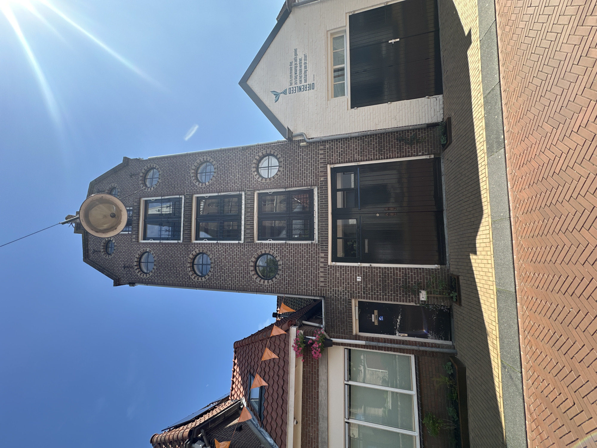The image shows a street scene in Steenwijk, Netherlands, on a sunny day. The focal point is a tall, narrow brick building with multiple circular windows and a large double door at street level. To the left of this building is a smaller brick building with a red-tiled roof adorned with orange triangular flags, and a large window. To the right of the tall building is a white building with a pitched roof, featuring a large black double door and text on the facade. The foreground is paved with red bricks arranged in a herringbone pattern, with lighter colored paving stones and a dark strip running parallel to the buildings. Shadows from the buildings are cast across the pavement, indicating the time of day. There are no people visible in the image.
