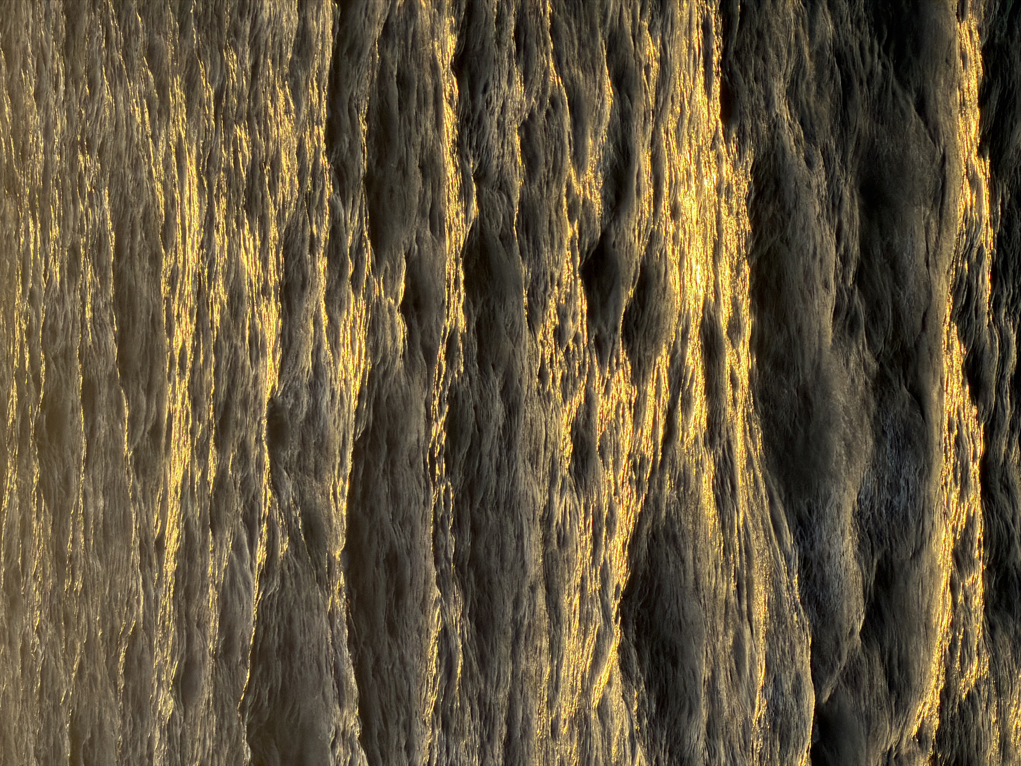 The image shows a textured surface with vertical patterns and variations in light and shadow. The color ranges from light gray to dark gray with a golden hue highlighting some of the raised areas. The texture appears rough and uneven. The location in the city of Unknown, Unknown is not determinable from the image. There are no people, subjects, objects, actions, or interactions visible. The environment is limited to the depicted textured surface.