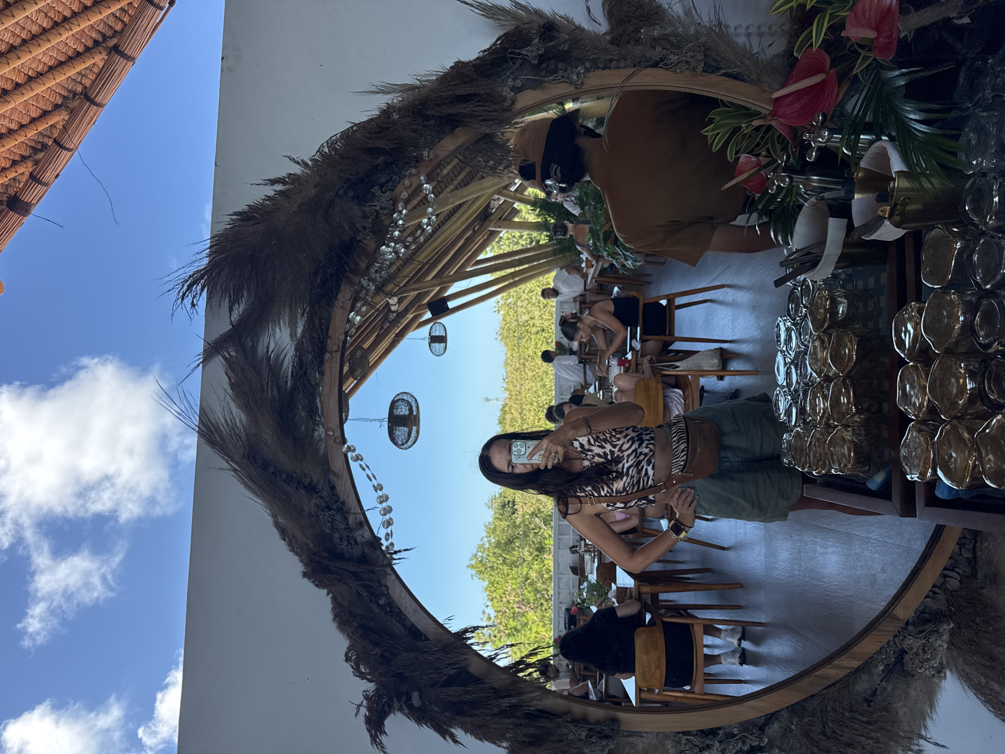 The image depicts a woman taking a selfie in a mirror at a restaurant in Kuta Selatan, Indonesia. The mirror is circular and decorated with dry grass and some crystals. Behind the woman, reflected in the mirror, are other patrons sitting at tables, and a bamboo structure. The restaurant's outdoor area is visible in the background. Some plants are placed near the mirror. Glasses are arranged on a shelf to the right. The outside sky can be seen in the mirror.