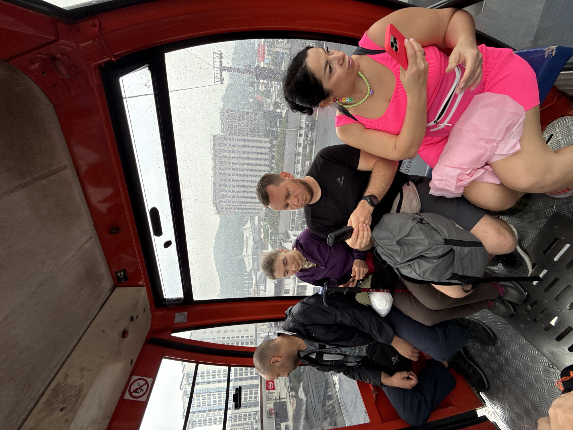Inside a red cable car ascending above Zhangjiajie, China, four people are seated. A woman in a bright pink outfit and colorful necklace holds a phone, looking towards the window. A man in a black shirt is beside her, looking down. A younger person in a purple shirt sits next to him with walking sticks. A fourth person in a black jacket sits at the bottom. Outside, rain streaks the windows, obscuring views of the city's buildings and landscape.