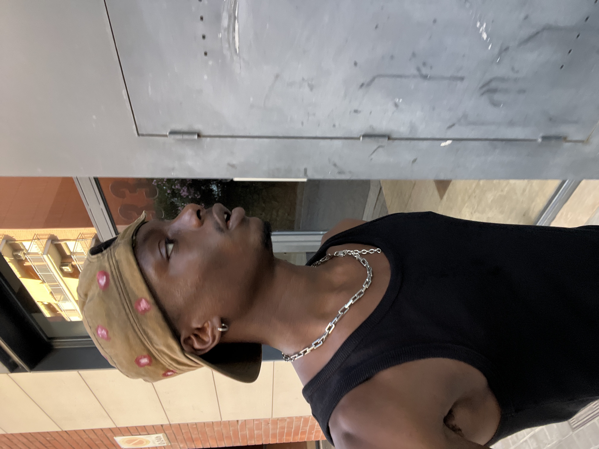 This vertical image features a young man of dark complexion, seen from the chest up, looking upwards and to his right, with his mouth slightly open. He is wearing a black sleeveless top, a silver-colored chain necklace with rectangular links, and a silver hoop earring in his left ear. On his head is a tan or light brown cap, decorated with multiple small, pink, possibly floral, patterns. He has a slight beard or stubble.
The setting appears to be an urban outdoor environment during the daytime, indicated by the bright lighting. Behind him, a large, light grey metal panel or door with visible hinges dominates the upper background. To his left, a large glass surface reflects a multi-story building with numerous windows and balconies, suggesting a city street or building exterior. The ground beneath him is a mix of light-colored paving and reddish-brown brickwork. A small, white rectangular sign with a red circular symbol (likely indicating 'no entry' or 'no parking') is partially visible on the ground, though any text on it is illegible. The man appears to be in a contemplative or observational state, possibly looking at something above or in the distance. The scene is consistent with an urban setting like Barcelona, Spain.