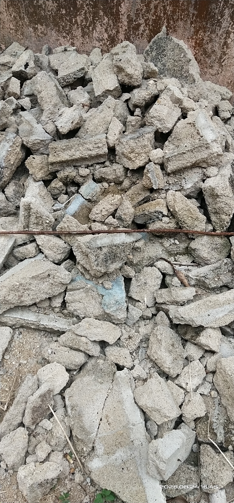 This is a close-up, eye-level, vertical shot of a large pile of broken concrete debris. The debris is rough and varied in size and shape, with some pieces appearing angular and sharp, while others are more rounded. The concrete ranges in color from light gray to a darker, more weathered gray, with some pieces showing hints of blue or white. In the upper portion of the image, a darker, textured surface is visible, which appears to be a wall or a similar structure. A thin, rusty metal rod or wire runs horizontally across the middle of the frame, separating the upper and lower sections of the debris pile. The ground beneath the pile is a mix of dirt and scattered smaller pieces of concrete, with a few dry grass stalks and some small green leaves emerging. There is no evidence of human activity, people, or specific landmarks that would indicate the city of Matabu, Indonesia. The timestamp in the lower right corner of the image reads "2026.08.24 16:45," which is a future date.