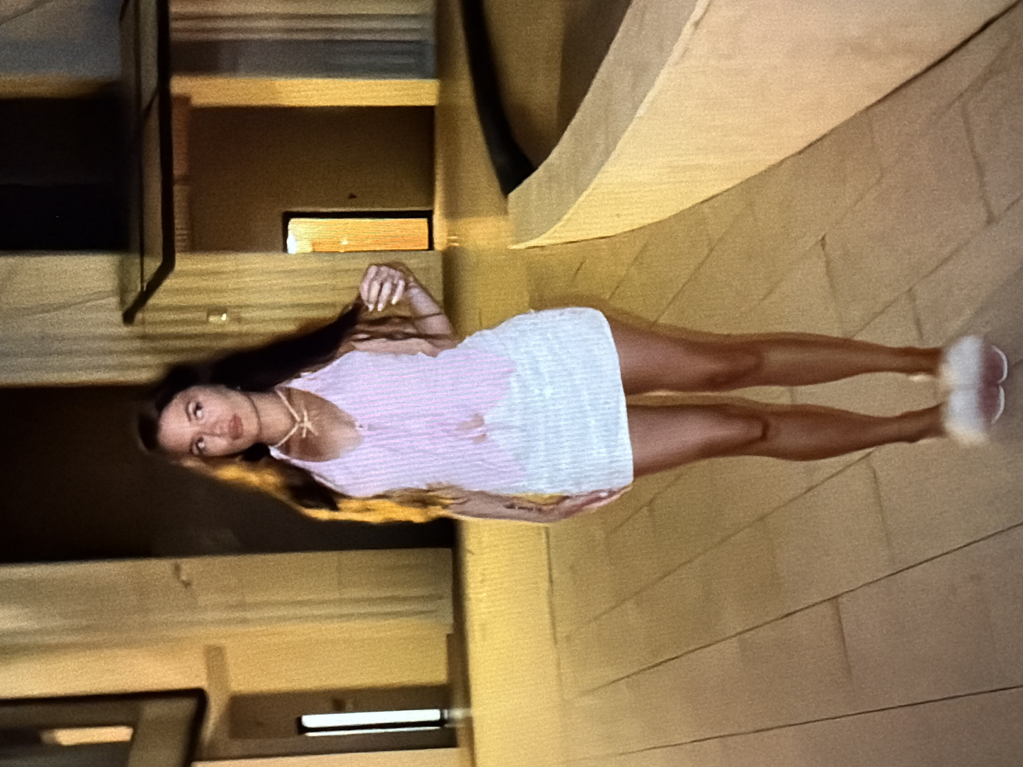 A full-body, medium-shot, slightly low-angle photograph captures a woman standing outdoors on a paved walkway. The woman has long, dark hair and is wearing a pink and white striped halter top with a plunging neckline and a white, textured mini-skirt. She is accessorized with a gold necklace. Her right hand is extended to touch her hair, and her left arm is by her side. She is wearing fluffy pink slippers. The background is blurred but suggests an urban environment with architectural elements and warm lighting. The ground is paved with rectangular tiles. There is a curved, light-colored structure to the right of the woman. The scene is brightly lit, with warm tones dominating the image. This scene is not located in Sirius, Russia.