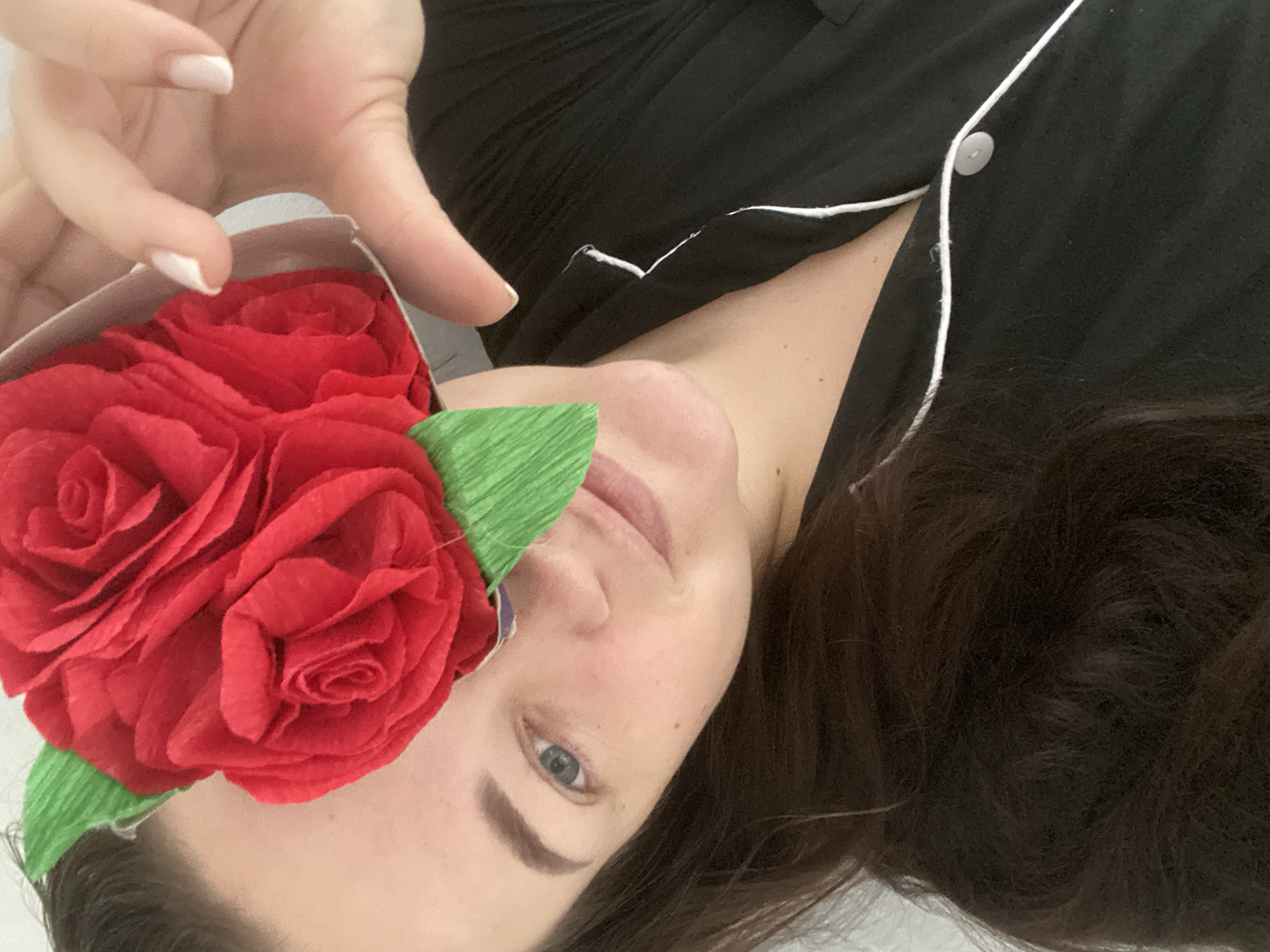 A person is lying down wearing a black pajama top with white piping and a single visible button, holding a white box filled with multiple red paper roses with green leaves partially covering their left eye. The person has dark brown hair and fair skin with visible freckles. The background is a plain white wall. There is no information available in the image to confirm the location is Sochi, Russia.