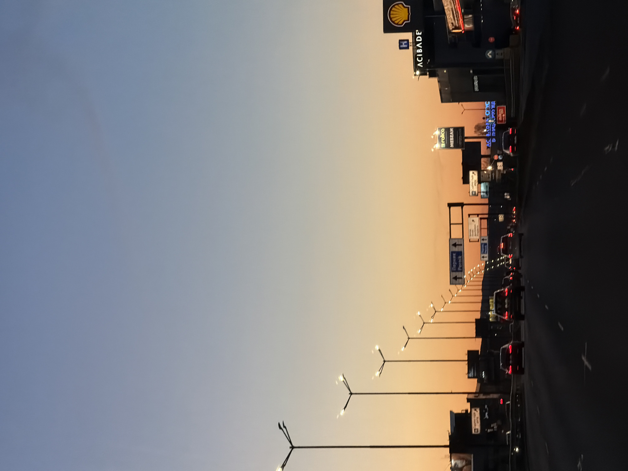 The image depicts a highway in Sofia, Bulgaria at dusk, with the sky transitioning from blue to orange. The Shell gas station sign is visible among other commercial signs. Cars are traveling along the highway, illuminated by streetlights lining the road. The scene includes light poles, traffic, and urban infrastructure.