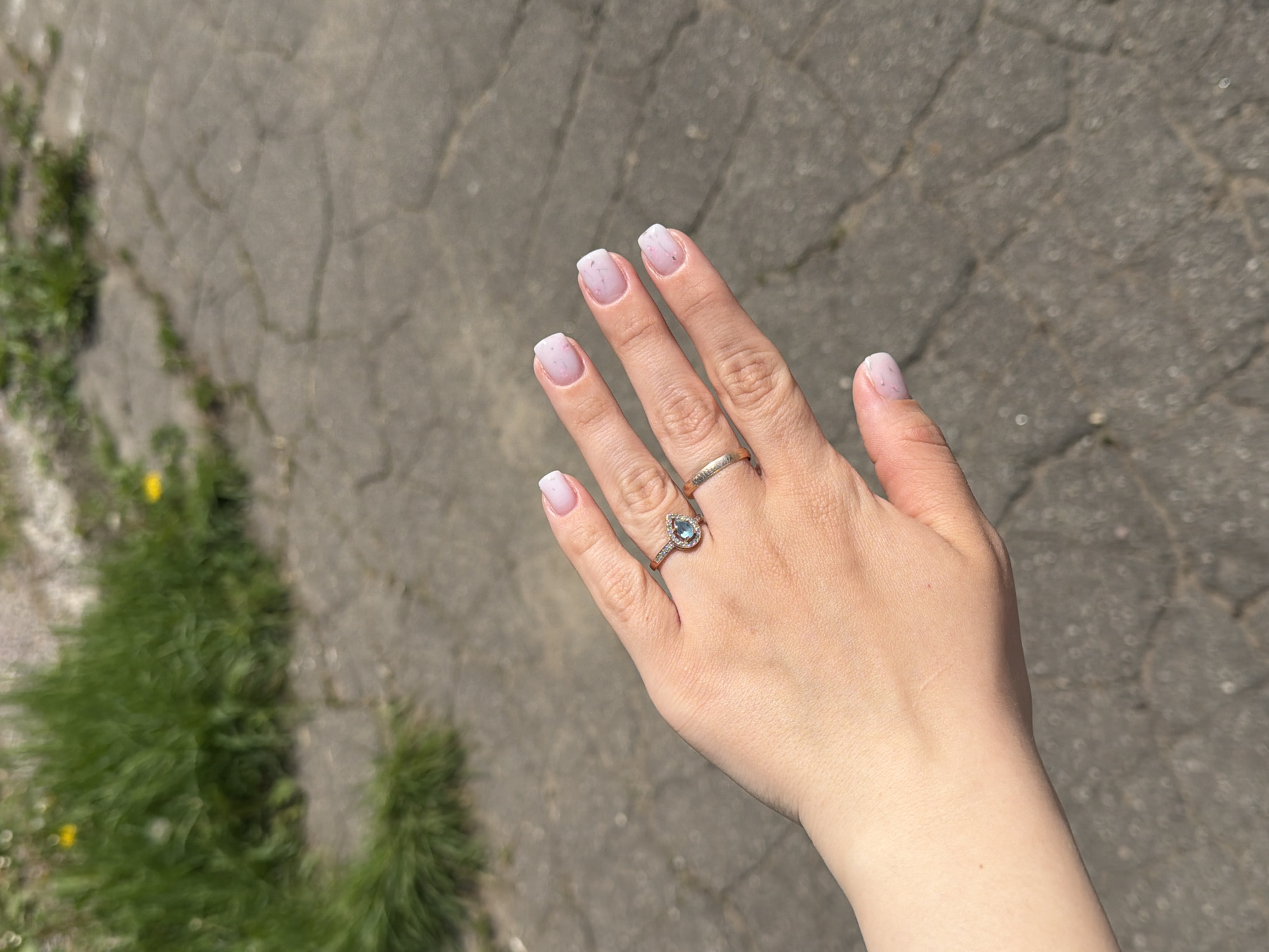 A woman's hand is extended, revealing manicured nails with a light pink base and white tips. The ring finger sports a gold band, while the middle finger features a gold ring with a pear-shaped, blue gemstone surrounded by smaller stones. The background consists of a paved surface with cracks and a patch of green foliage to the left. The scene appears to be outdoors, possibly a sidewalk or patio. There is no evidence to confirm this image was taken in Reshetnikovo, Russia.