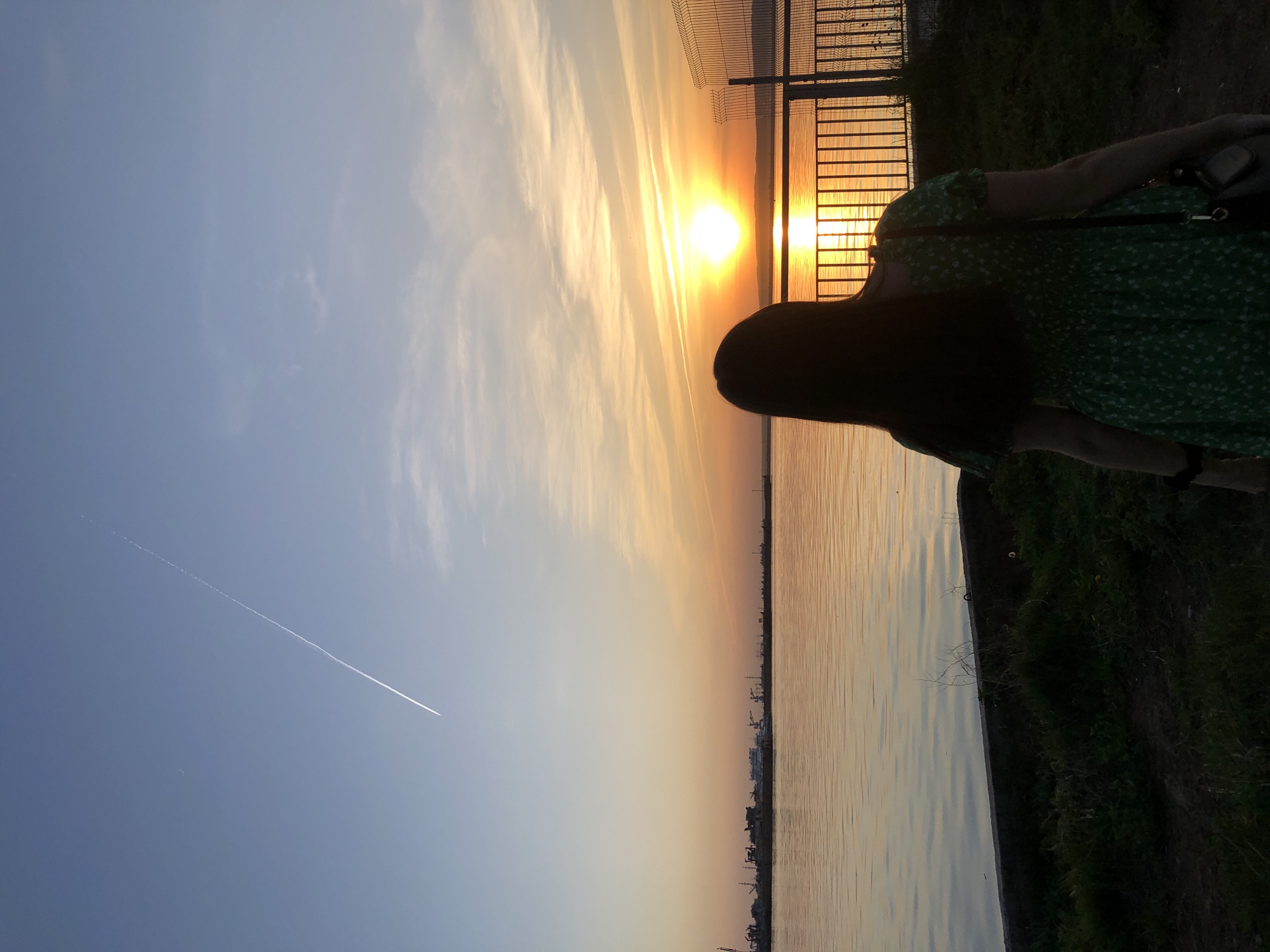 A woman stands near a body of water at sunset in the city of Chaikovsky, Russia. She is wearing a green dress and has long, dark hair. A metal fence runs parallel to the water's edge. The sky is a mix of blue and orange, with a contrail visible in the blue portion. The water reflects the colors of the sky. On the opposite bank, a cityscape is visible. The foreground includes a grassy area.