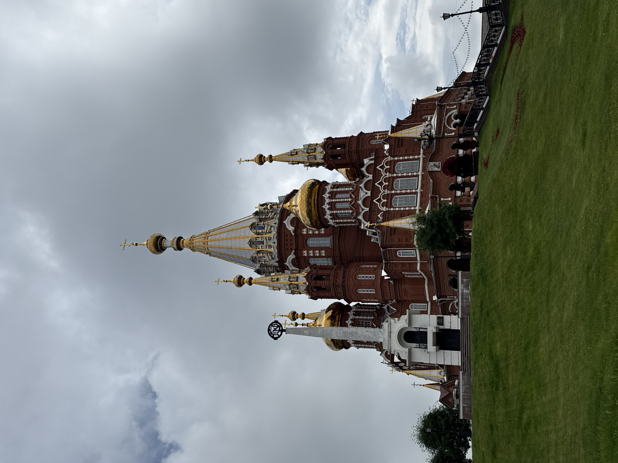 The image shows the Ascension Cathedral, a prominent landmark in Izhevsk, Russia. The cathedral is a large, red-brick Orthodox church with multiple golden domes and a tall, ornate spire. It is situated on a grassy hill, with a white monument in front of it, adorned with a dark circular emblem. The sky is overcast with dramatic clouds. Stairs lead up to the entrance of the cathedral, and decorative lights are strung along the railing. Trees are visible on the left side of the image, and a lamppost stands near the stairs. The scene appears to be set during the daytime.