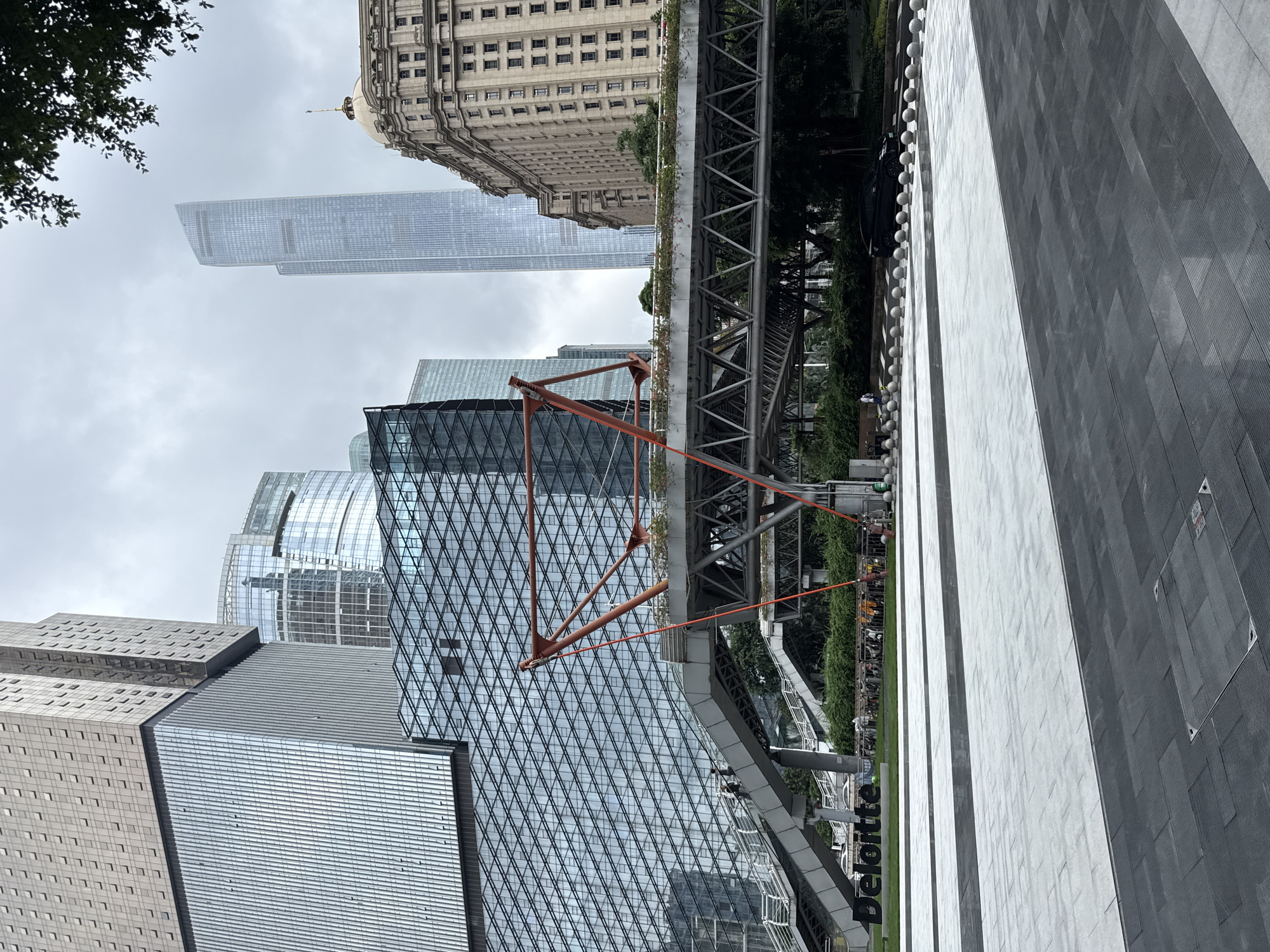 In Tianhe District, China, an outdoor plaza scene unfolds, dominated by modern architecture and an elevated walkway. A large, multi-story building with a glass facade and metallic accents stands prominently on the left. Adjacent to it, a complex structure with a geometric glass pattern and a bright orange, angular support system forms a distinctive pedestrian bridge. This walkway is lined with greenery and connects to elevated staircases. In the foreground, a paved area transitions from light grey to dark grey tiles, with a manhole cover visible in the darker section. The "Deloitte" logo is displayed in large, black block letters on the grass in front of the building. In the background, several skyscrapers pierce the overcast sky, including a very tall, slender glass tower. A traditional-looking, ornate building with a dome is visible on the far right. A dark car is parked on the street behind a row of bollards. Several people are present, some walking on the elevated walkway and stairs, and others near the "Deloitte" sign. A bicycle rack is also visible. The overall impression is one of a contemporary urban environment.