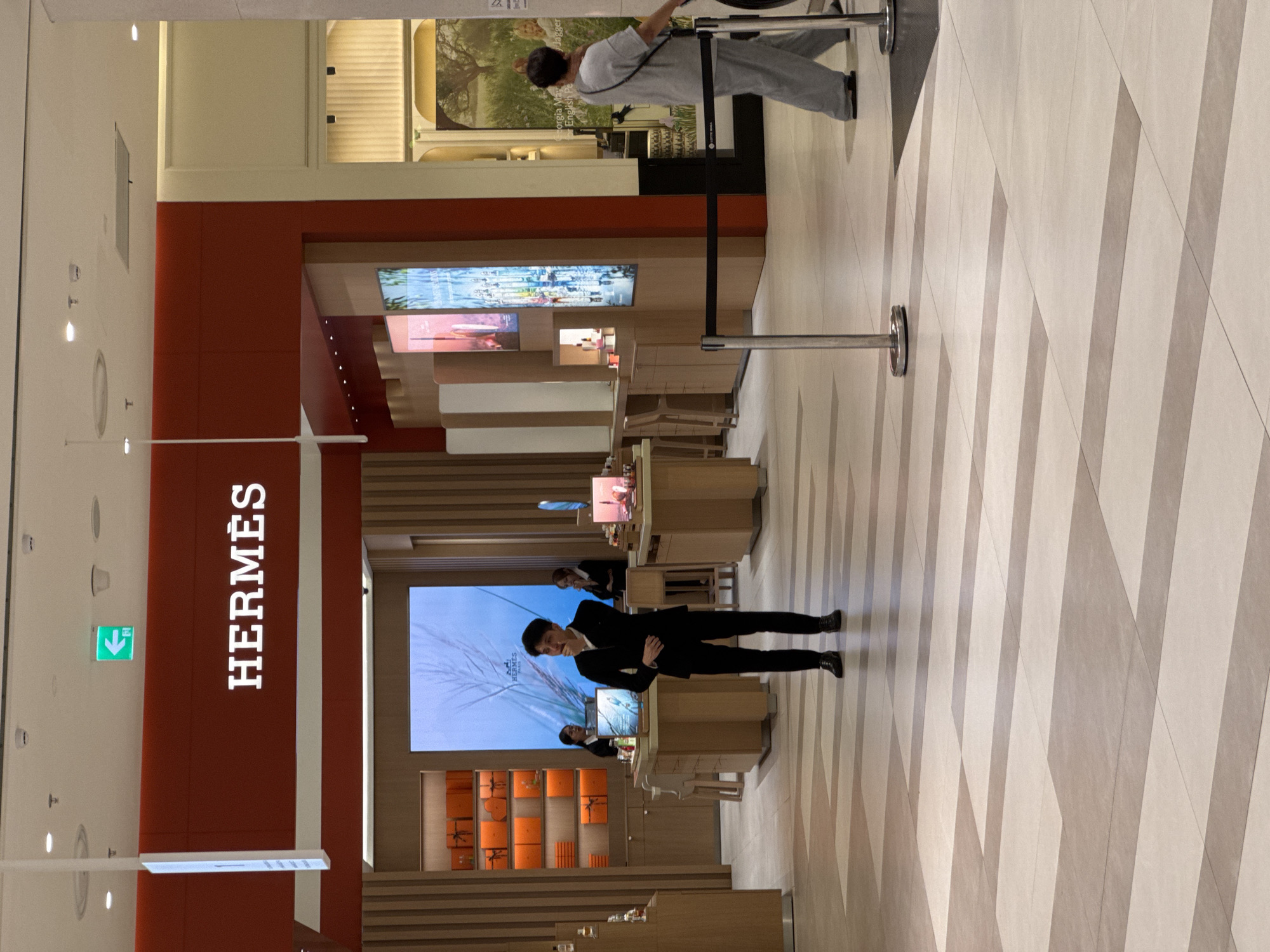 The image captures the interior of a high-end retail store, likely a luxury brand boutique, with a focus on the "Hermès" branding. A large red pillar prominently displays the "HERMÈS" logo in white capital letters.
In the foreground, a man dressed in a black suit is striking a pose, with his left hand on his hip and his right arm bent, looking towards the camera. He appears to be an employee or model. Behind him, a large digital screen displays the Hermès logo above an image of wheat stalks. On shelves below, numerous orange boxes, a signature Hermès packaging, are neatly arranged.
Further into the store, the architecture features clean lines and a warm, inviting color palette, with wooden accents and a patterned tiled floor. Another person, wearing a light grey uniform, is visible in the background near the entrance, stretching their arms upwards.
The setting suggests an indoor environment, possibly a mall or a standalone boutique, given the polished floors and architectural design. The lighting indicates it could be daytime, but the artificial lighting of the store is prominent. No weather cues are visible. The overall mood is one of sophistication and luxury, conveyed by the branding, the merchandise display, and the attentive presentation of the staff. The visible text includes the "HERMÈS" logo on the pillar and the screen, as well as a green emergency exit sign with an arrow.