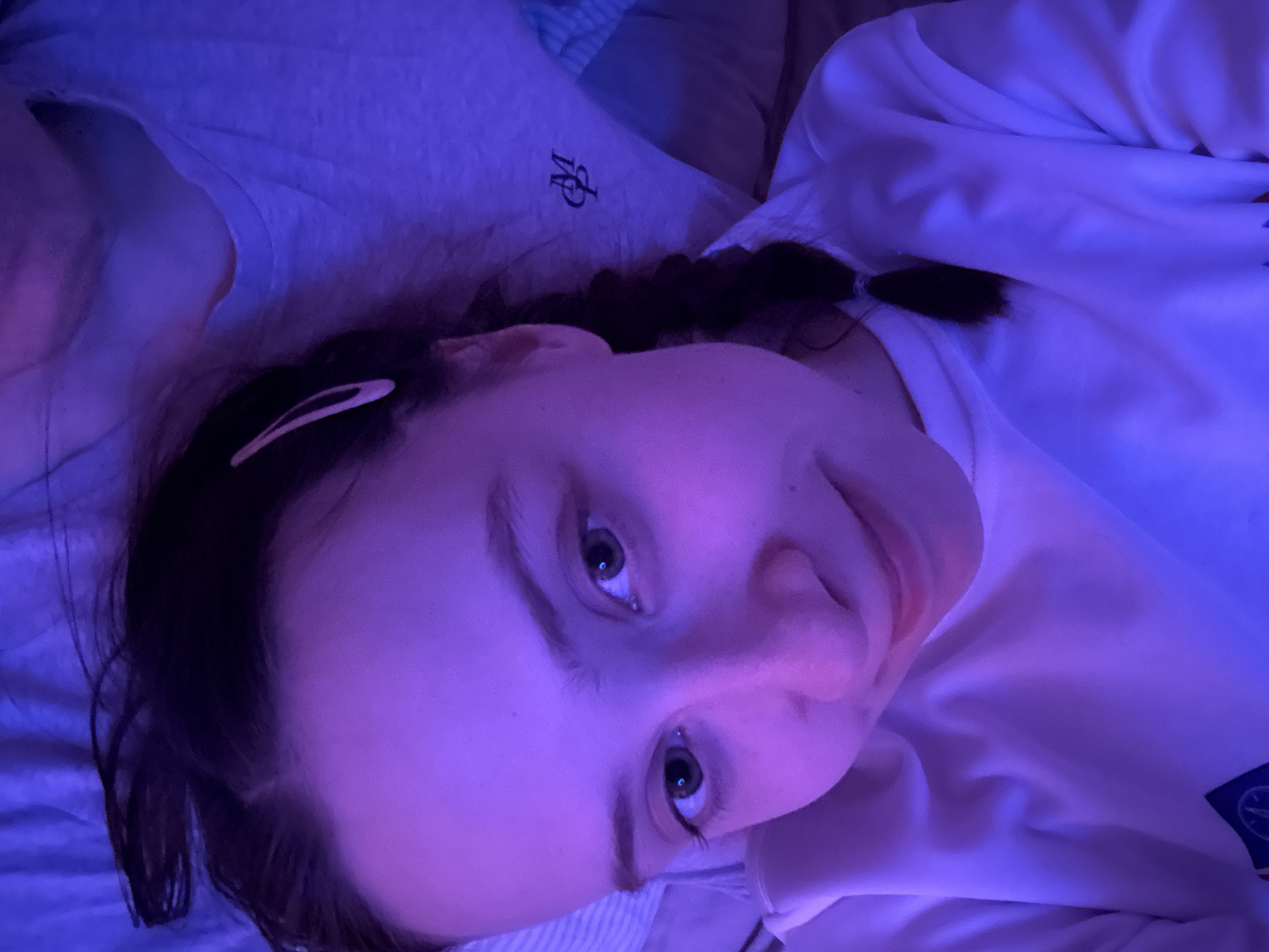 A woman lies on a bed in a room illuminated by blue light. She has dark hair pulled back with a pink hair clip and a single braid. She is wearing a white long-sleeved shirt with a logo on the front. A gray shirt with a monogram is partially visible behind her. The striped bedding is also visible. The location of Прикамский, Russia, cannot be determined from the image.