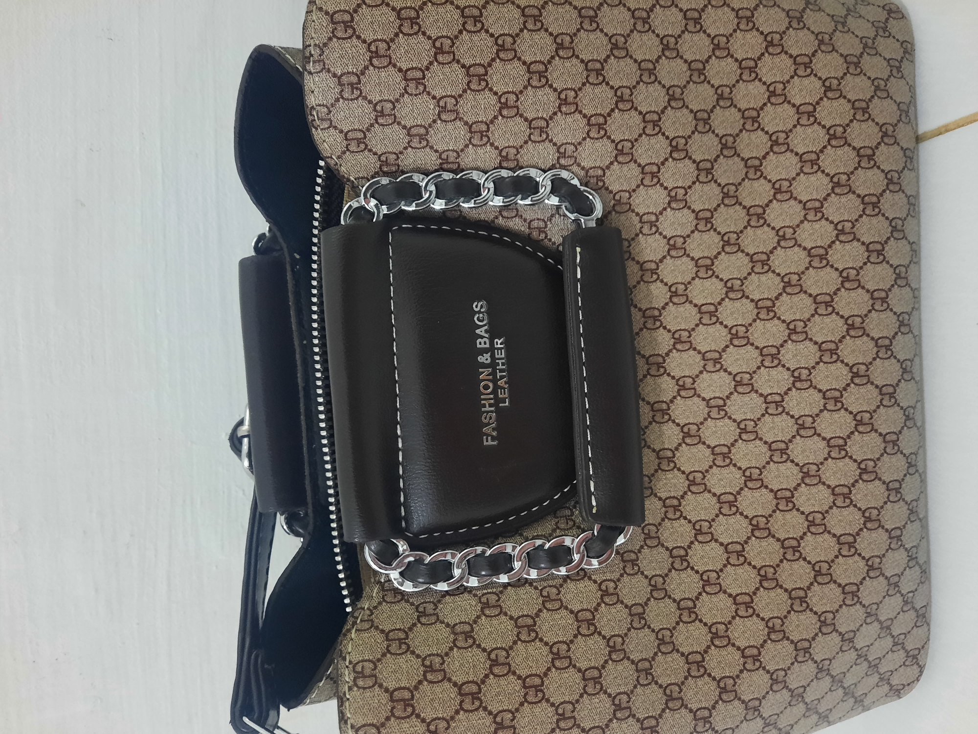 A brown and tan patterned handbag sits on a white surface. The handbag has a brown leather trim with white stitching, and a silver chain detail. The pattern on the handbag appears to be a repeating "GD" logo. Text on the brown leather trim reads "FASHION & BAGS LEATHER". The background is a white, textured wall. The lighting is neutral, suggesting an indoor setting. The image is cropped to focus on the handbag. No people or specific location details within Lhokseumawe, Indonesia are visible.