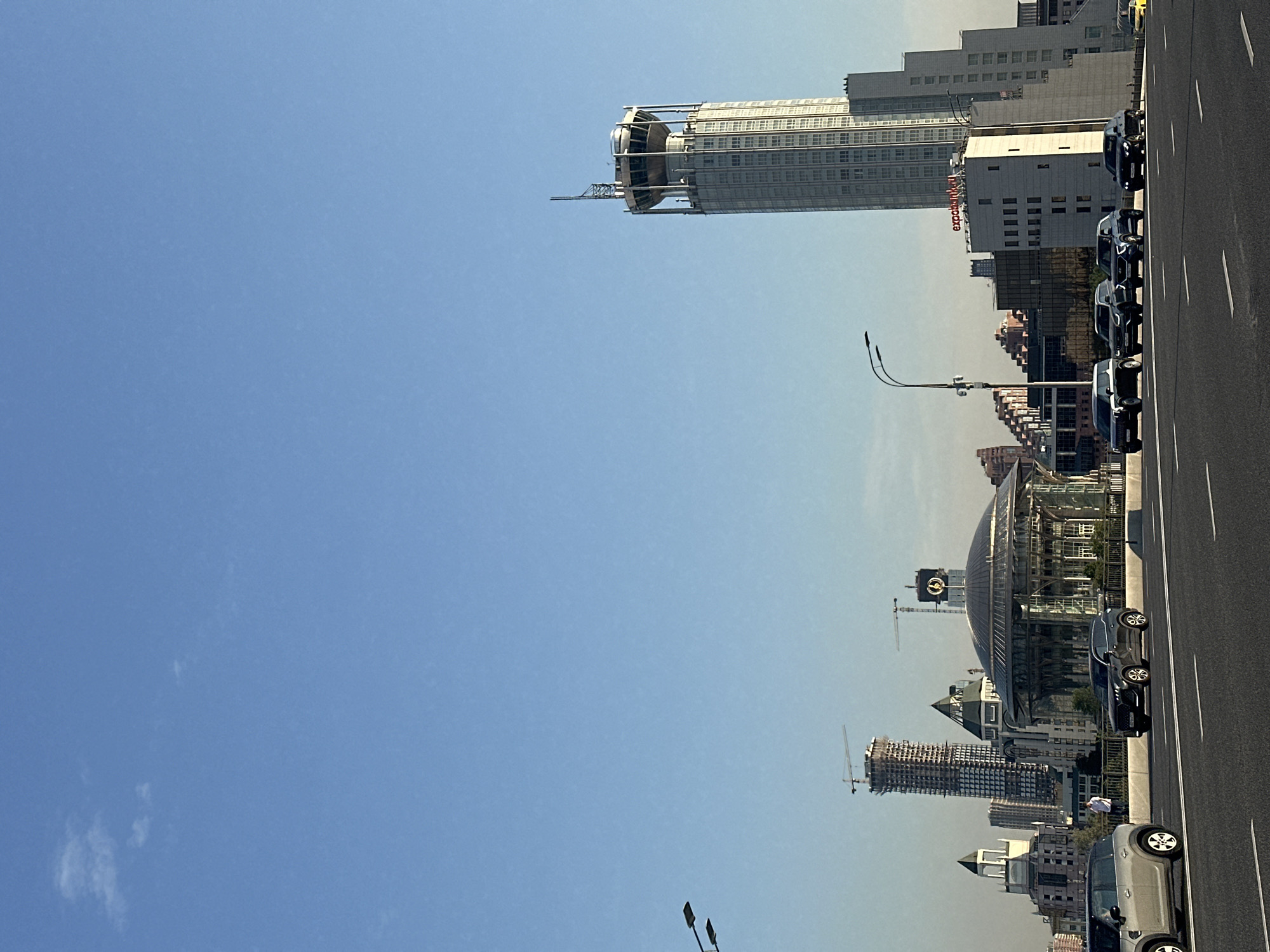 The image shows a cityscape in Moscow, Russia, likely taken from a bridge or elevated roadway. The foreground features a multi-lane asphalt road with white lane markings. Several cars are driving on the road, including a silver SUV on the left, a dark SUV, and a black sedan. In the middle ground, there is a large, modern building with a distinctive domed glass roof, resembling a concert hall or theater. Behind this structure, several other buildings are visible, including a very tall, cylindrical skyscraper with a distinctive circular platform at its top, adorned with antennas and a railing. Another skyscraper under construction, with visible scaffolding and cranes, is also present. The background is dominated by a clear, light blue sky with a few wispy clouds. The overall scene depicts a bustling urban environment with a mix of contemporary and developing architecture under a bright sky.