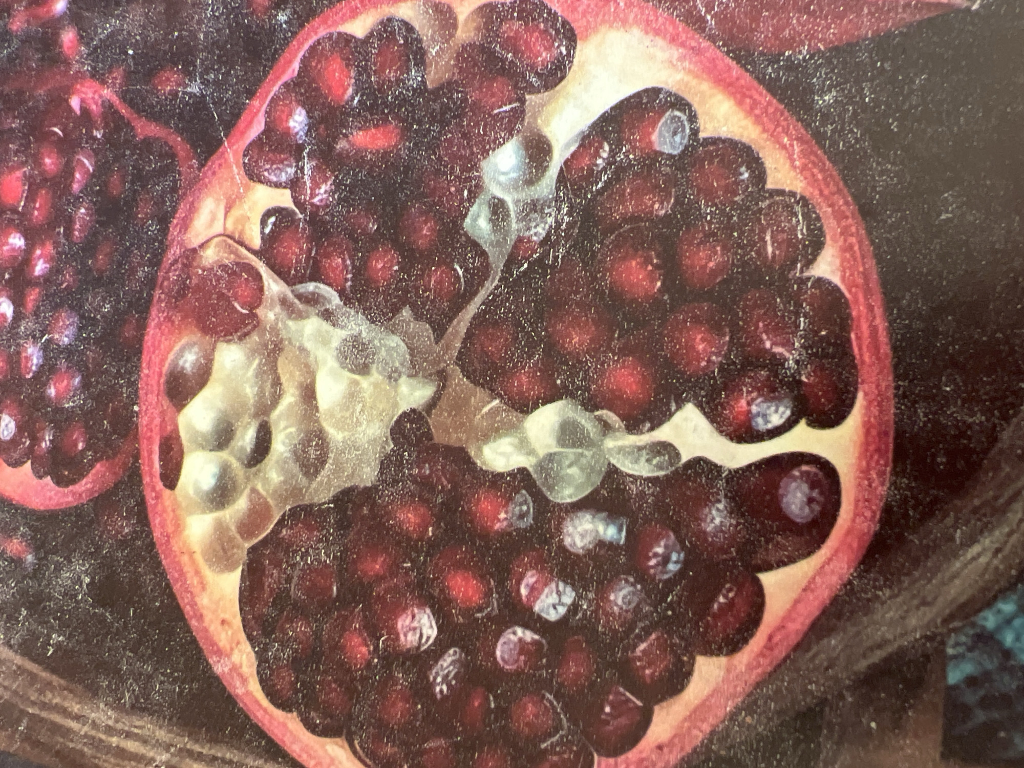 The image shows a close-up of a halved pomegranate. The arils, which are the edible seeds, are a deep red color, and some are partially transparent, revealing the seeds within. White membranes separate the arils. The pomegranate rind is a vibrant pinkish-red. The background is dark and textured, possibly representing a dark cloth or surface. There are no people or any other identifiable objects or settings visible in the image. There is no information within the image that would allow for the determination of its location in Minsk, Belarus.