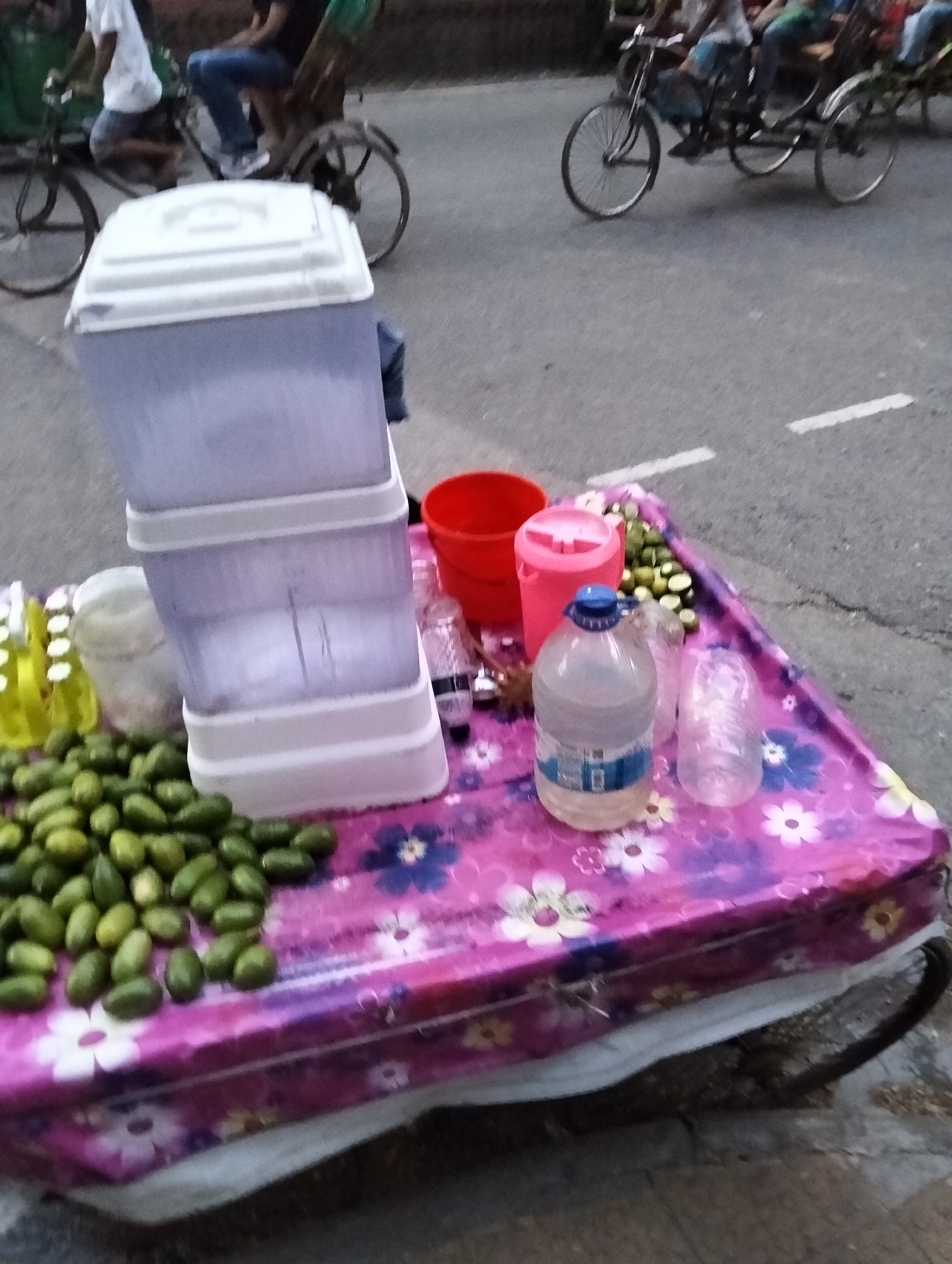 A street vendor's mobile stand is in Dhaka, Bangladesh. The stand, likely a modified rickshaw, is covered with a pink floral tablecloth. On it are drinks in a layered dispenser, a red bucket, bottles, plastic cups, and a pile of small green fruits, possibly olives or unripe mangoes. In the background, people are riding bicycle rickshaws along the street.