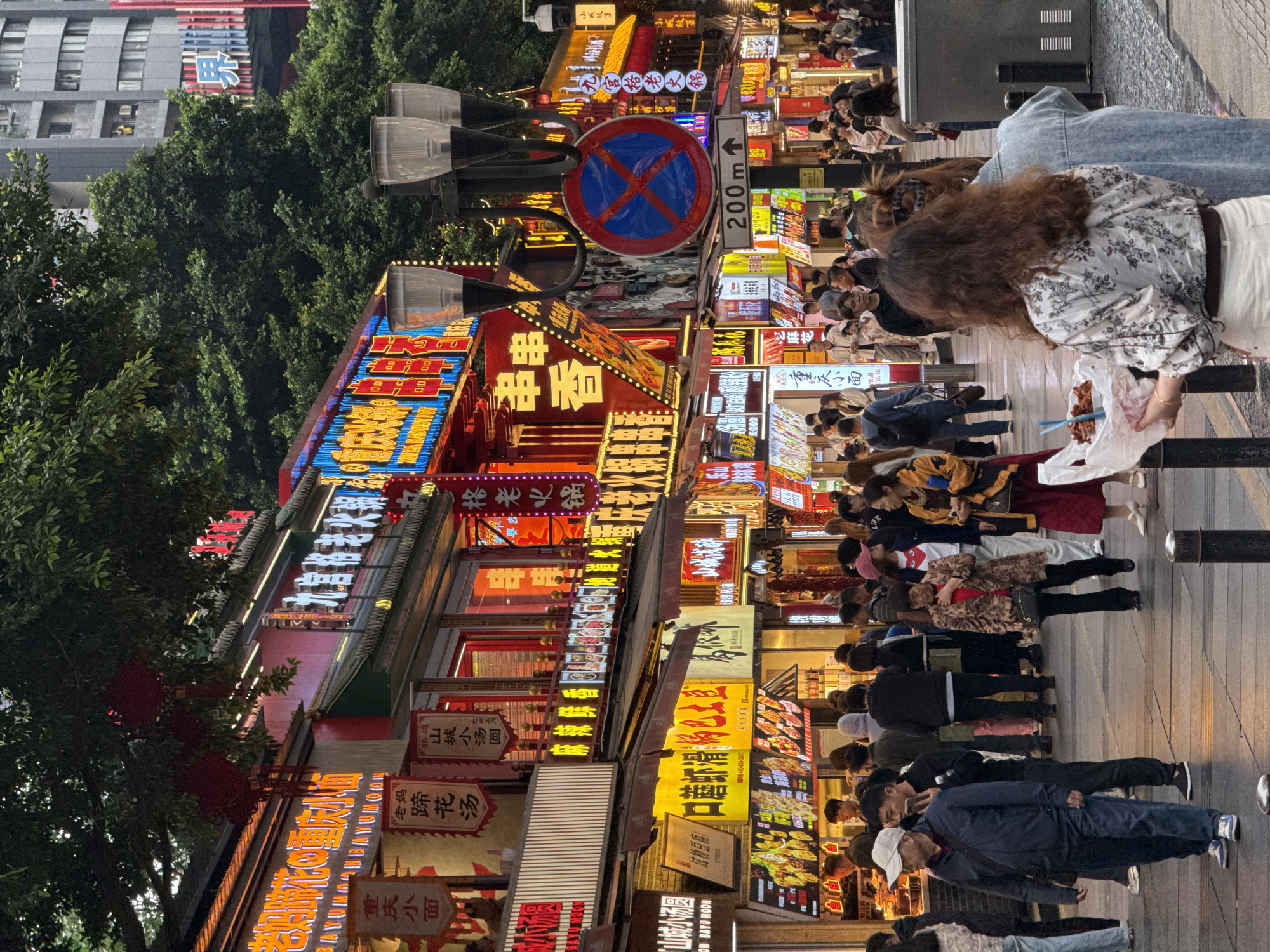 The image captures a bustling street scene in Yuzhong District, Chongqing, China, illuminated by a myriad of brightly lit storefront signs. Numerous pedestrians are walking along the sidewalk, some engaged with food in hand. The signs display various Chinese characters and restaurant names, adding to the vibrant atmosphere. Trees flank the left side of the street, while tall buildings rise in the background. A traffic sign indicating "200m" and "no parking" is visible above the shops. The overall impression is of a busy commercial area at night.