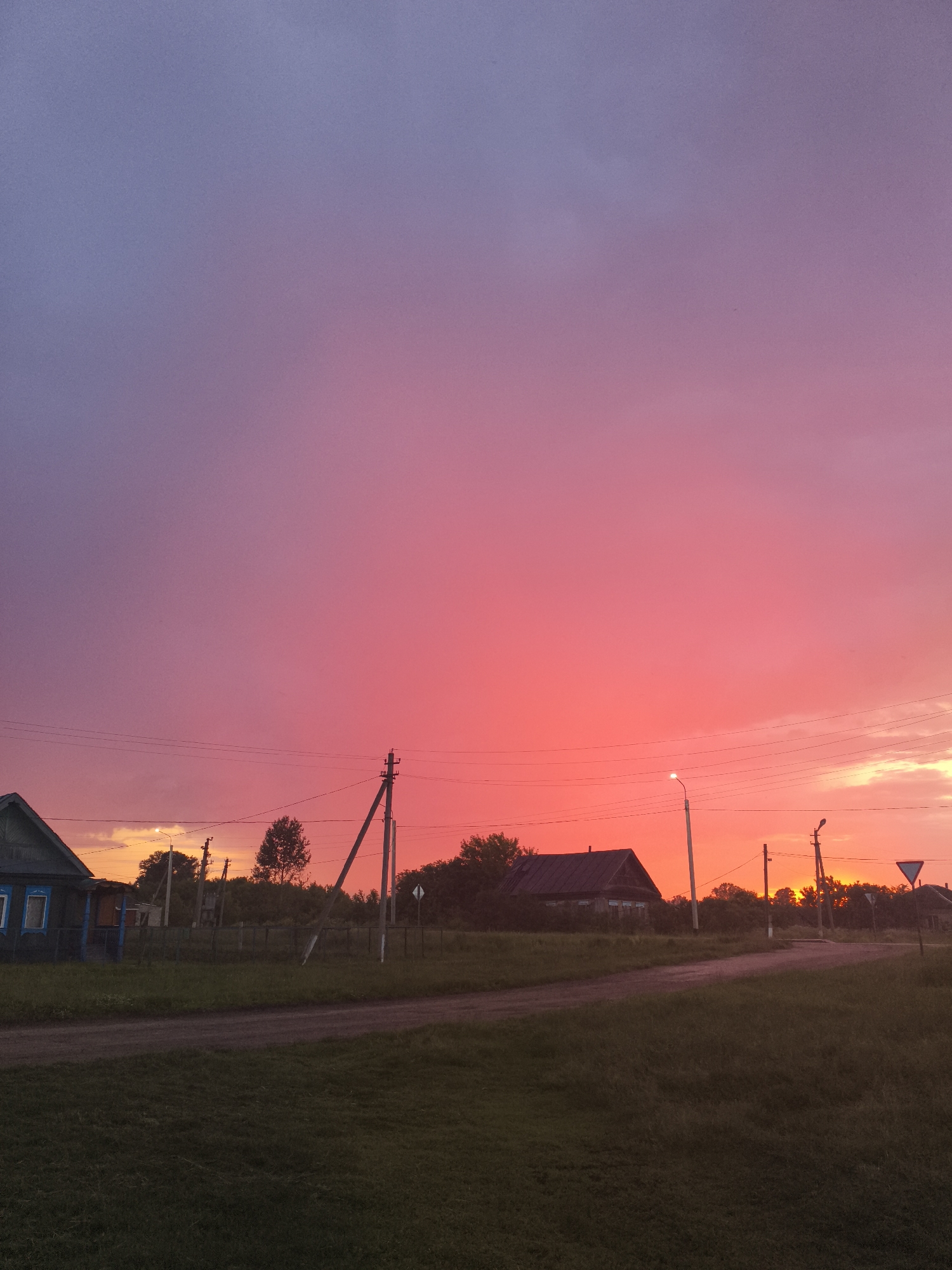 A wide, low-angle shot shows a rural road in the village of Ilmino, Russia, under a dramatic sunset sky. The sky transitions from a deep violet at the top to a vibrant pink and then to a fiery orange near the horizon. The road, a dirt path, curves from the foreground to the middle ground. On the left side of the road, a small, dark wooden house with blue-trimmed windows is visible. Beyond the road, silhouetted against the bright sky, are several power poles, trees, and a few other dark, indistinct structures. The ground in the foreground and middle ground is covered with grass. The overall impression is of a quiet, rural evening.
