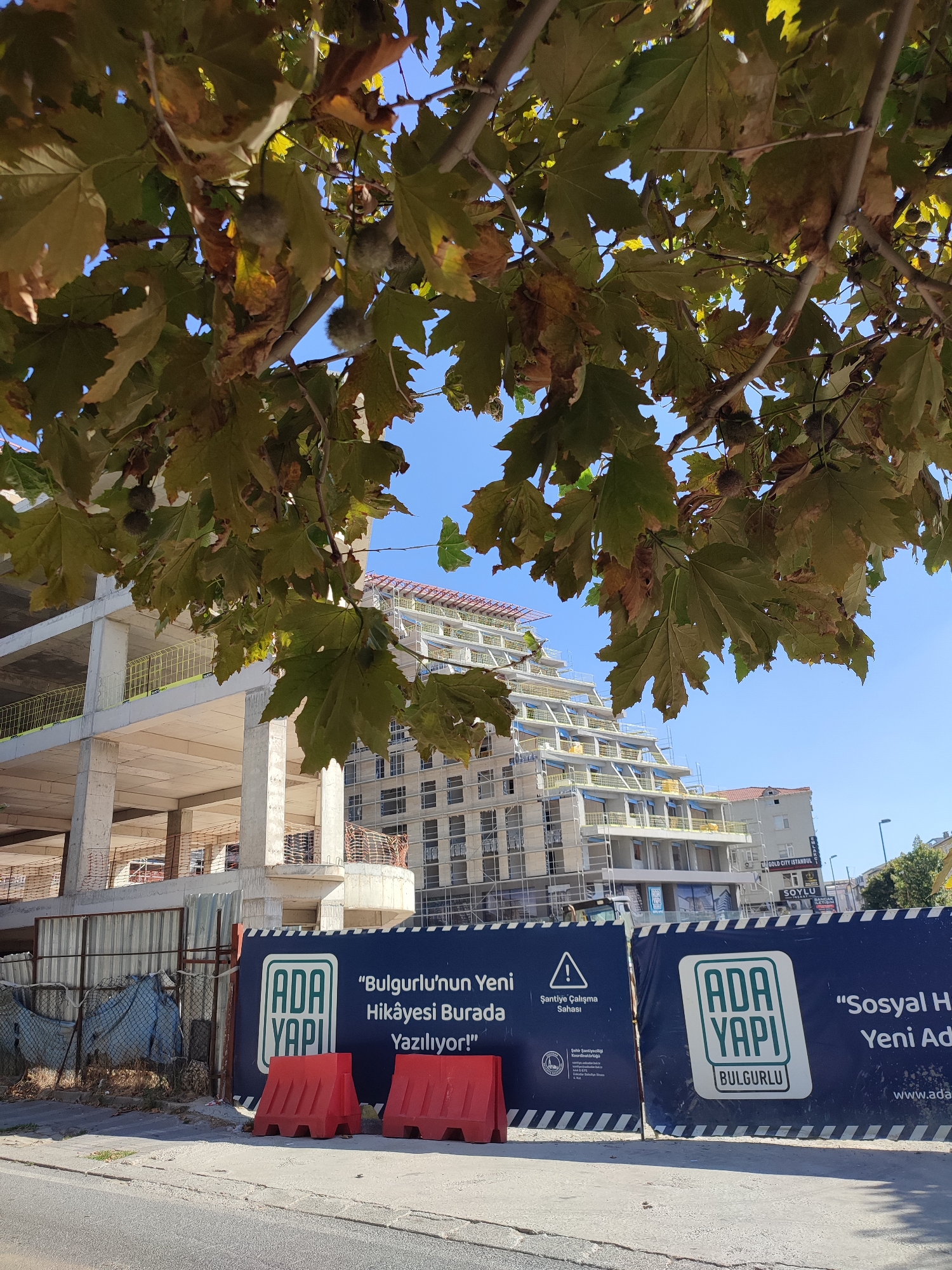 In Üsküdar, Turkey, a construction site is visible behind a fence adorned with banners. One banner displays the logo "ADA YAPI" in green and white, with the text "BULGURLU" underneath. Another banner features the same logo and the slogan "Bulgurlu'nun Yeni Hikâyesi Burada Yazılıyor!" which translates to "Bulgurlu's New Story is Written Here!". A third banner has the text "Sosyal Hayatin Yeni Adresi" meaning "New Address of Social Life". The construction site features a building in progress with multiple levels and scaffolding. In the foreground, two red plastic barriers are placed on the asphalt. Overhanging the scene are the branches and large leaves of a tree, some of which are showing signs of autumn colors. The sky is clear and blue.