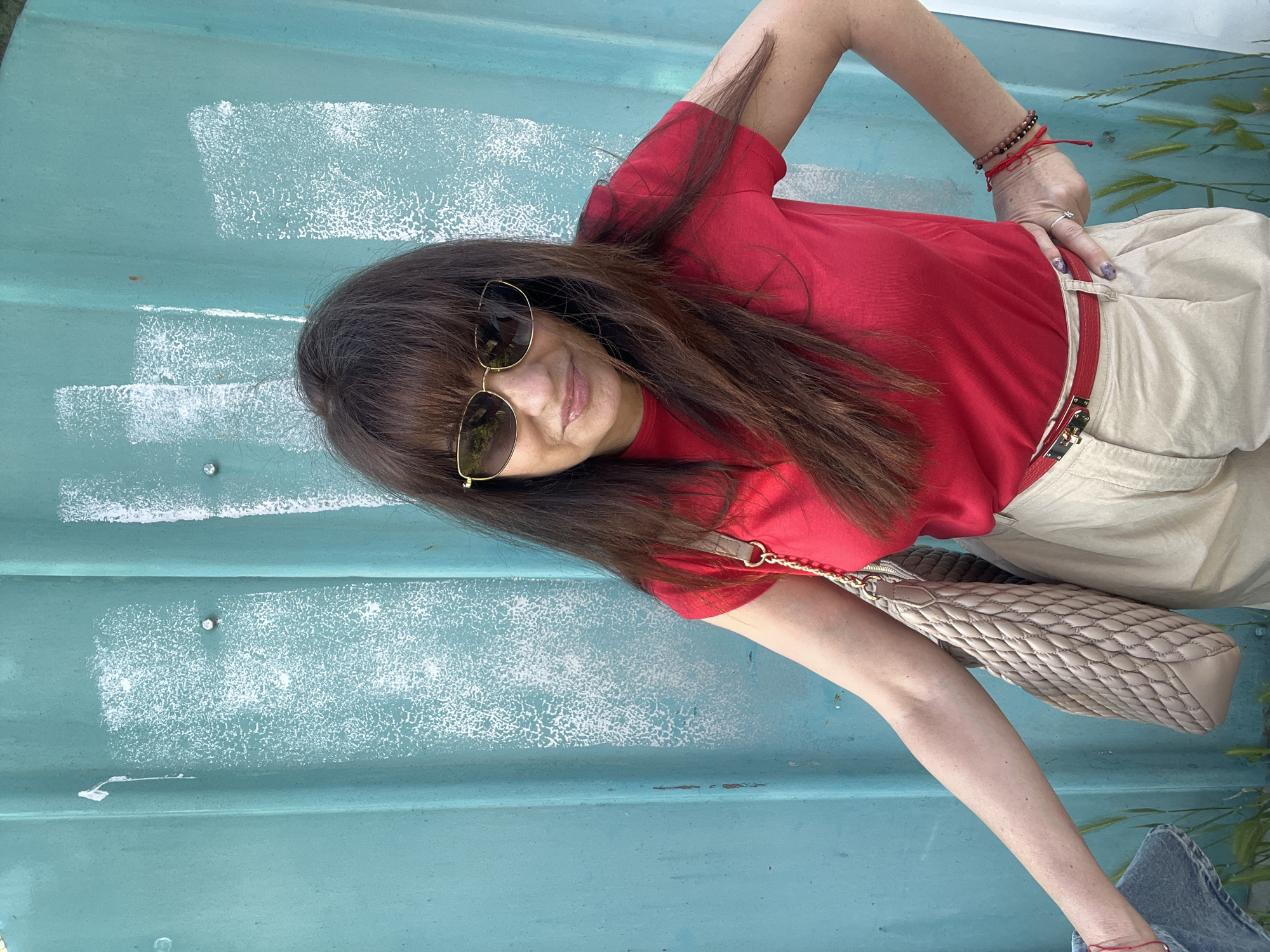 A woman in a red t-shirt and khaki pants, wearing sunglasses, is positioned against a turquoise-painted wall. She has a beige quilted purse hanging from her shoulder. Red string bracelets are visible on her wrists. The wall behind her features remnants of white paint in rectangular shapes. The location is outdoors, but the specific place in Plovdiv, Bulgaria, cannot be determined from the image.