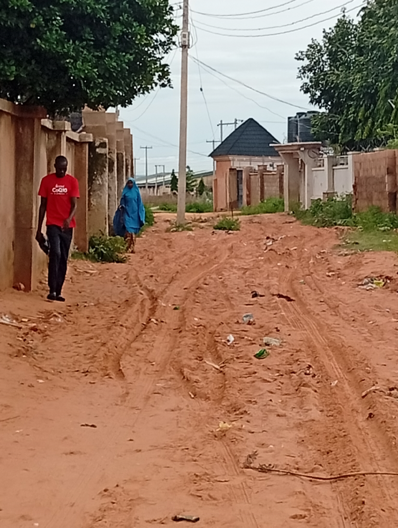 In Tumfure, Nigeria, two individuals walk along a dirt road. A man in a red t-shirt and dark pants walks on the left, holding a black object in his right hand. To his right and further down the road, a person in a blue hijab walks. The road is unpaved, with tire tracks visible, and littered with various pieces of debris. Walls line the left side of the road, with trees and greenery visible beyond them. Buildings and more vegetation are present in the background, along with utility poles and overhead wires. The sky is overcast.