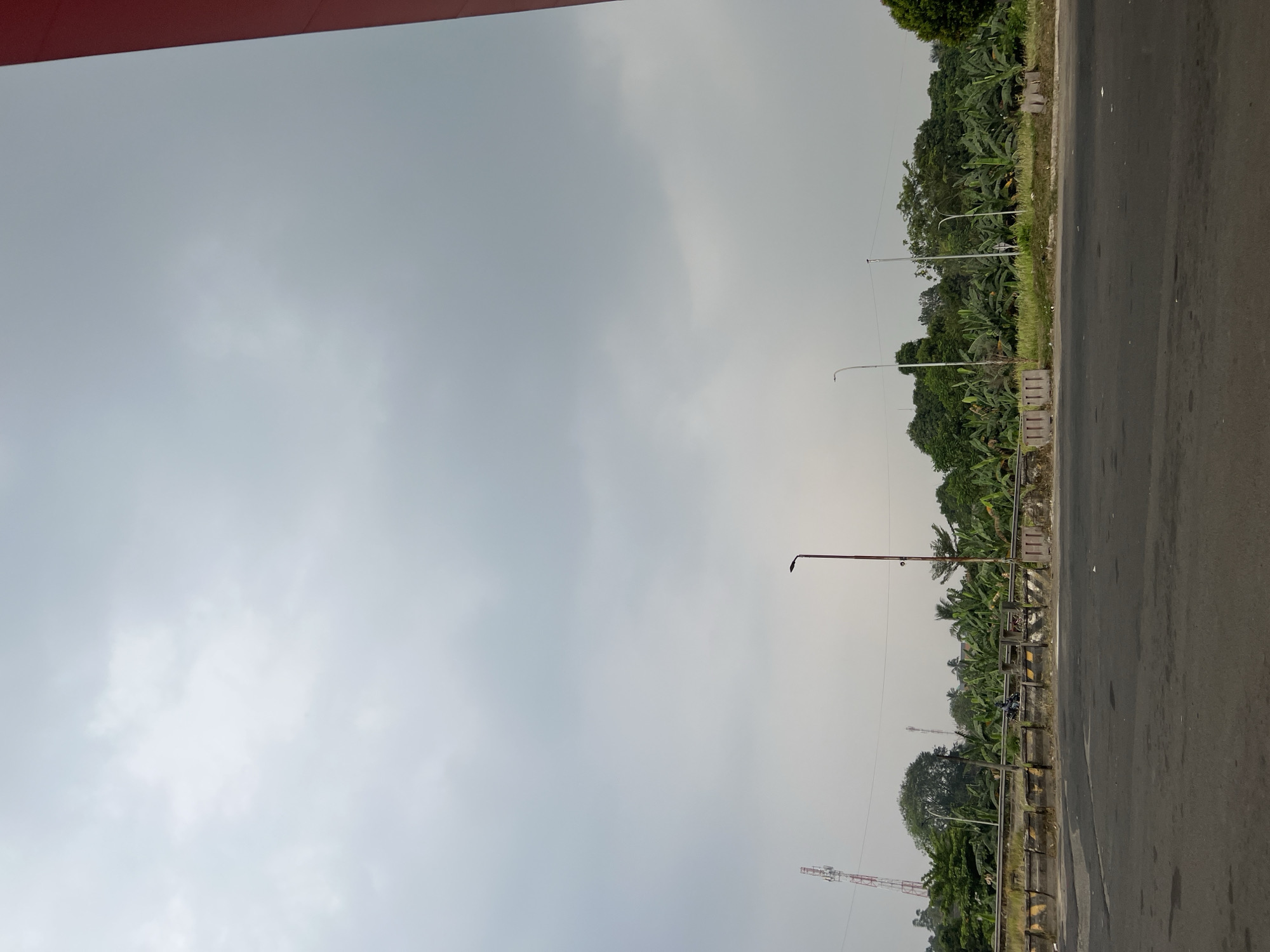 A wide-angle, medium-height shot captures a view of a road and a line of lush green trees with banana plants in the foreground. The sky is overcast, with a diffused, pale gray light. In the background, a tall communication tower with red and white markings stands to the left, and a lamppost is visible in the center. The road in the foreground appears to be asphalt, with some scuff marks and debris. A concrete barrier with yellow and black markings separates the road from the vegetated area. There is a motorcycle with a rider parked near the barrier. The overall scene suggests an outdoor environment in a semi-urban or rural area. The image is devoid of any specific landmarks that would pinpoint its exact location within Gunung Putri, Indonesia.