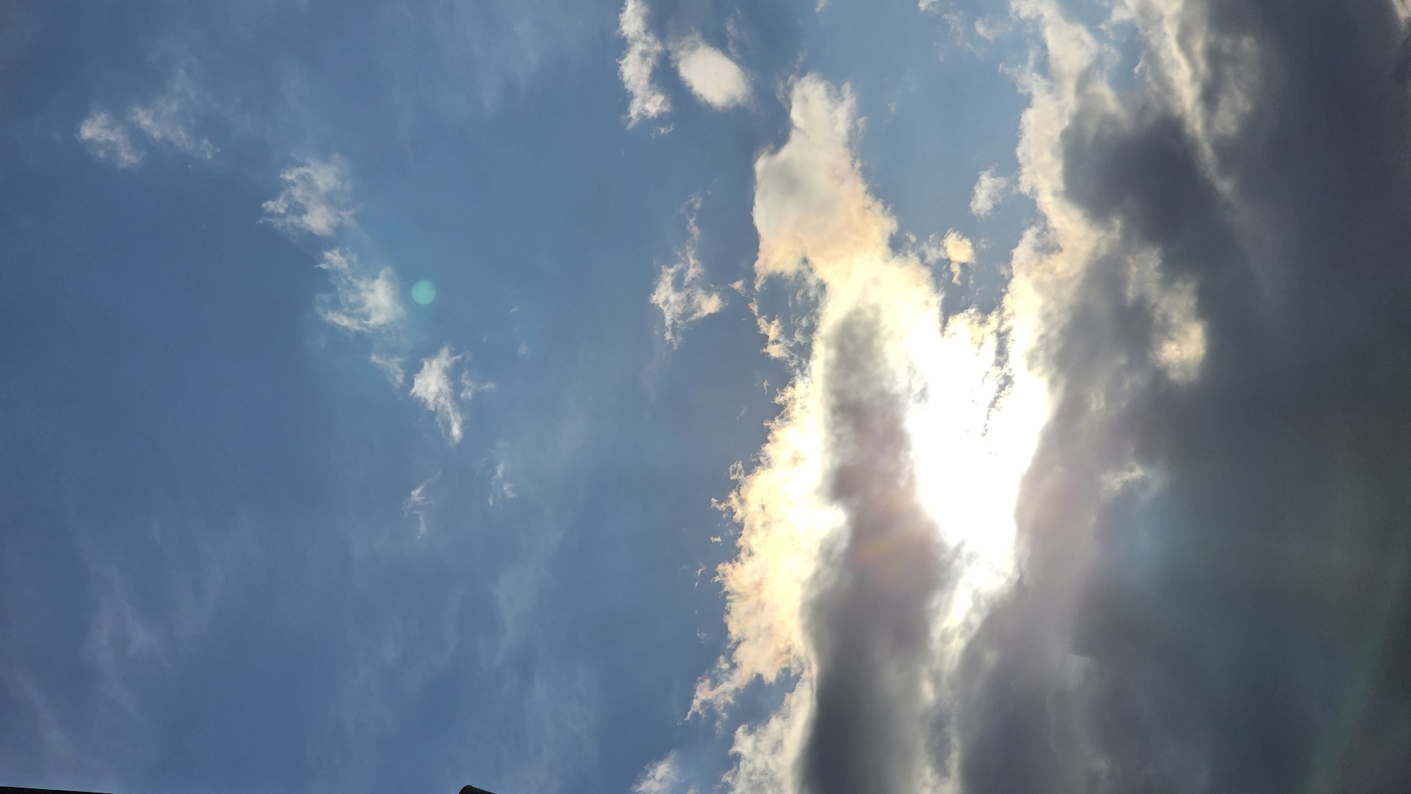 The image depicts a daytime sky with a mix of clouds and blue sky. Some clouds appear thin and white, while others are darker and denser, partially obscuring the sun. The sunlight is visible through the clouds, creating a bright, diffused light. The overall perspective is looking upwards. There is no identifiable information that points to the location as the city of Blagoevgrad, Bulgaria. No people, subjects, objects, actions, or interactions are visible other than the cloudscape itself.