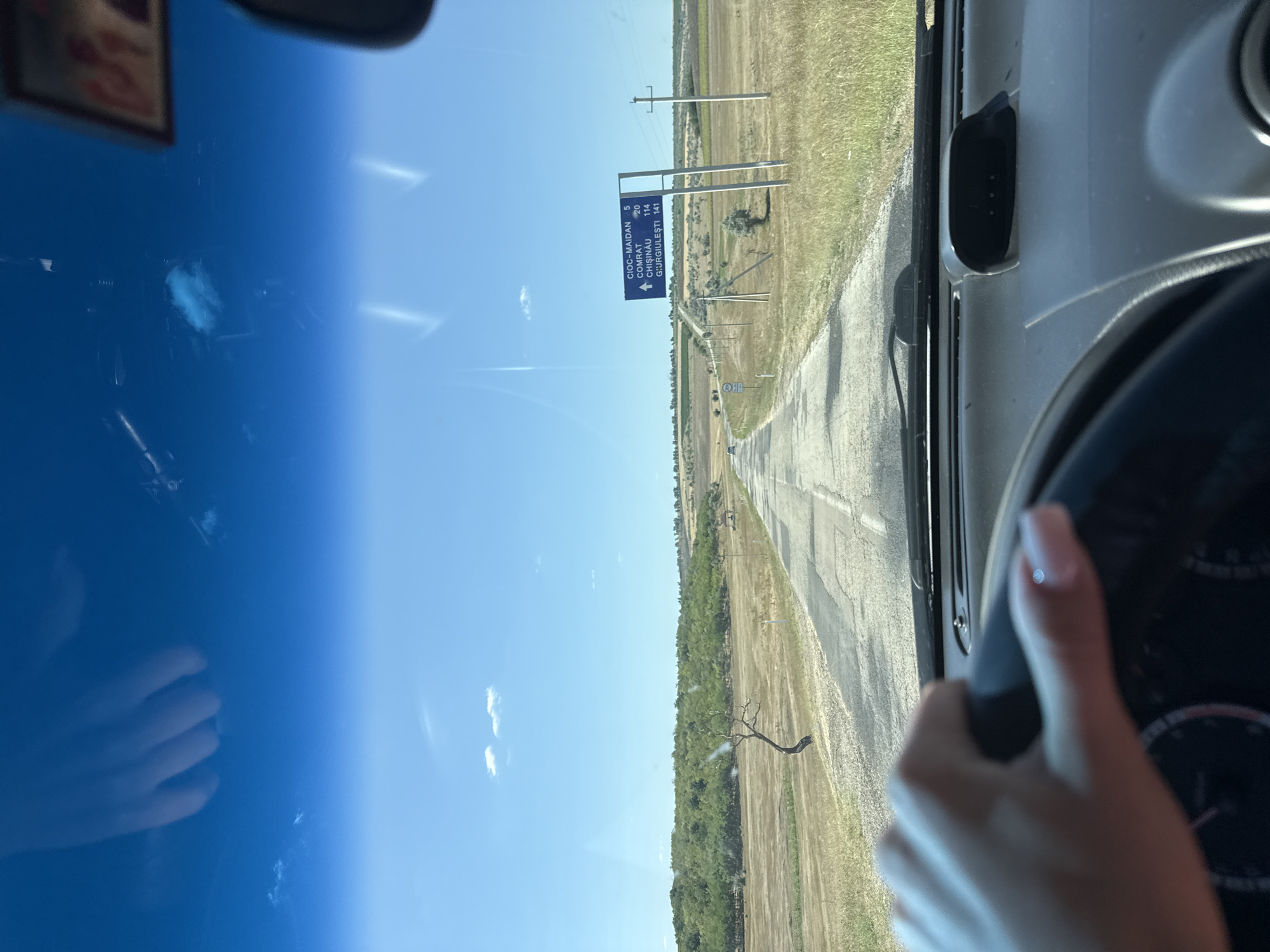The image is taken from the interior of a vehicle, looking forward through the windshield. A hand with manicured fingernails is visible on the steering wheel. The road ahead is asphalt, cracked and uneven, stretching into the distance through a dry, grassy landscape with scattered trees and bushes. Telephone poles line the right side of the road. In the distance, a blue road sign indicates directions and distances to "CIOC-MAIDAN 5", "COMRA T 20", "CHISINAU 114", and "GIURGIULESTI 141". The sky is bright blue with a few scattered white clouds. The environment suggests a rural, possibly agricultural area. The presence of the road sign and the distances listed point to this location being in the vicinity of Basarabeasca, Moldova, as it lists common destinations in that region.