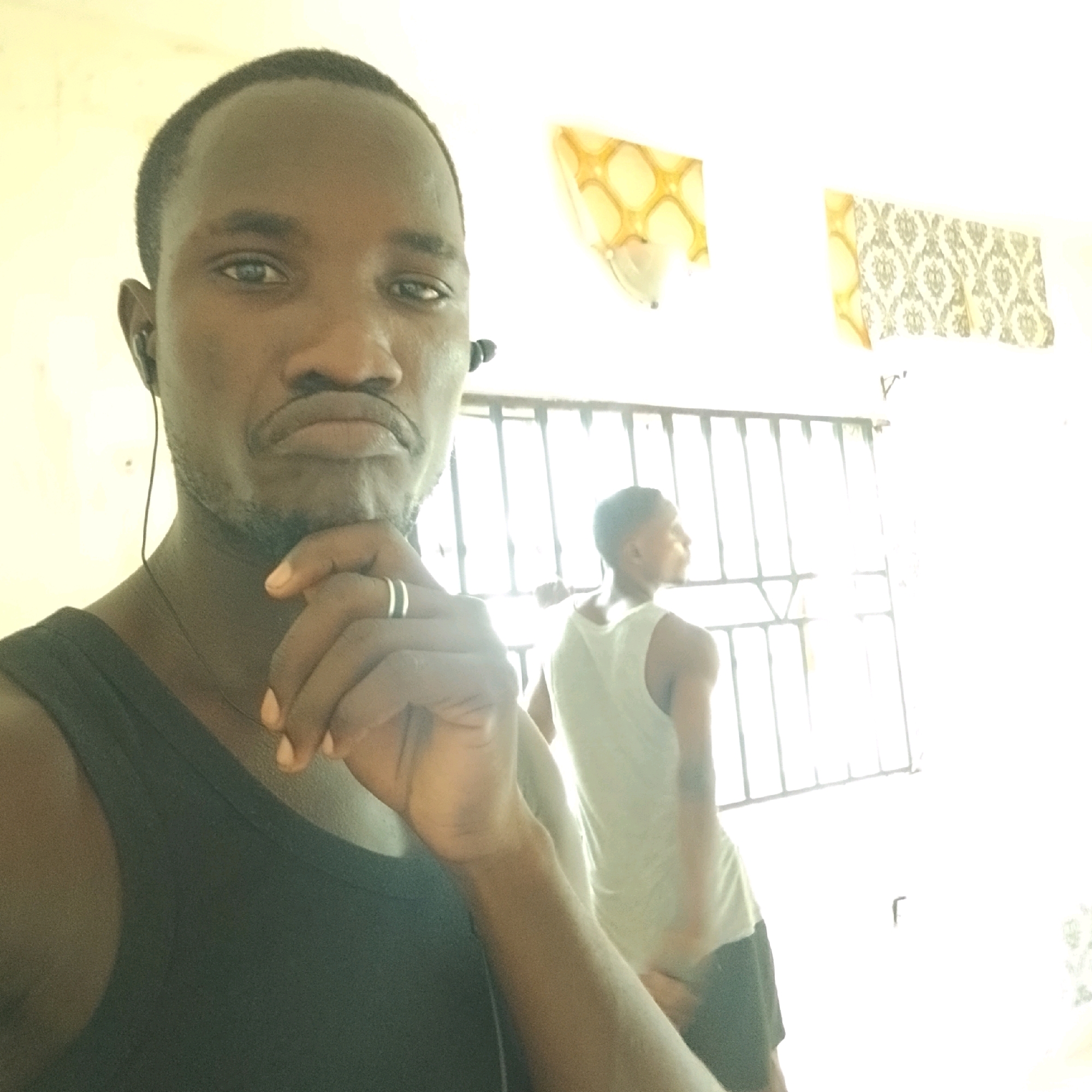 The image shows a close-up selfie of a Black male in the foreground, wearing a black tank top and earphones. He has short dark hair, a mustache, and a goatee. His left hand is raised, with his index finger touching his chin and his thumb supporting his jaw. A silver ring with two bands is visible on his left ring finger.
In the background, a second Black male, also wearing a light-colored tank top and dark shorts, is visible from the chest down, facing away from the camera. He appears to be standing in front of a window with horizontal bars.
The environment appears to be indoors, with a white wall and a light-colored ceiling. Two decorative light fixtures or wall hangings with patterned designs are visible on the wall in the background. The lighting is bright, with a yellowish hue, suggesting it might be natural light.
The presence of bars on the window could suggest a room with security features, possibly a detention center or a secure facility. Given the details of the people, clothing, and environment, it is possible this image was taken in Ado, Nigeria, although specific landmarks or identifying features of the city are not present in the image.